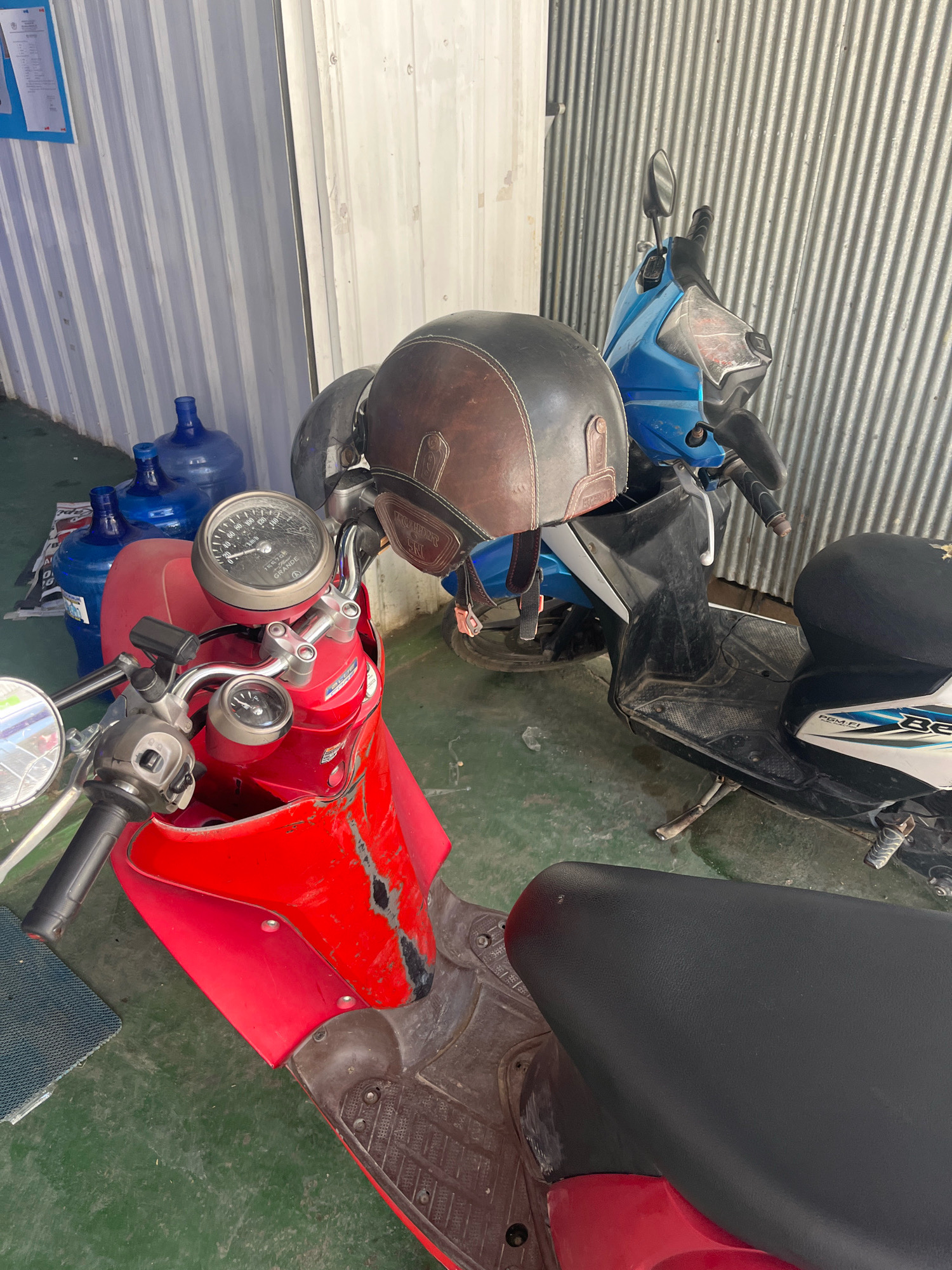 In Sengkang, Indonesia, two scooters are parked side-by-side against a corrugated metal wall. The scooter on the left is bright red and heavily scratched, with a visible speedometer and a brown helmet resting on its handlebars. Next to it, a blue scooter is partially obscured, its handlebars and a black helmet also visible. In the background, several large blue water jugs are stacked. The ground is a dull green color.
