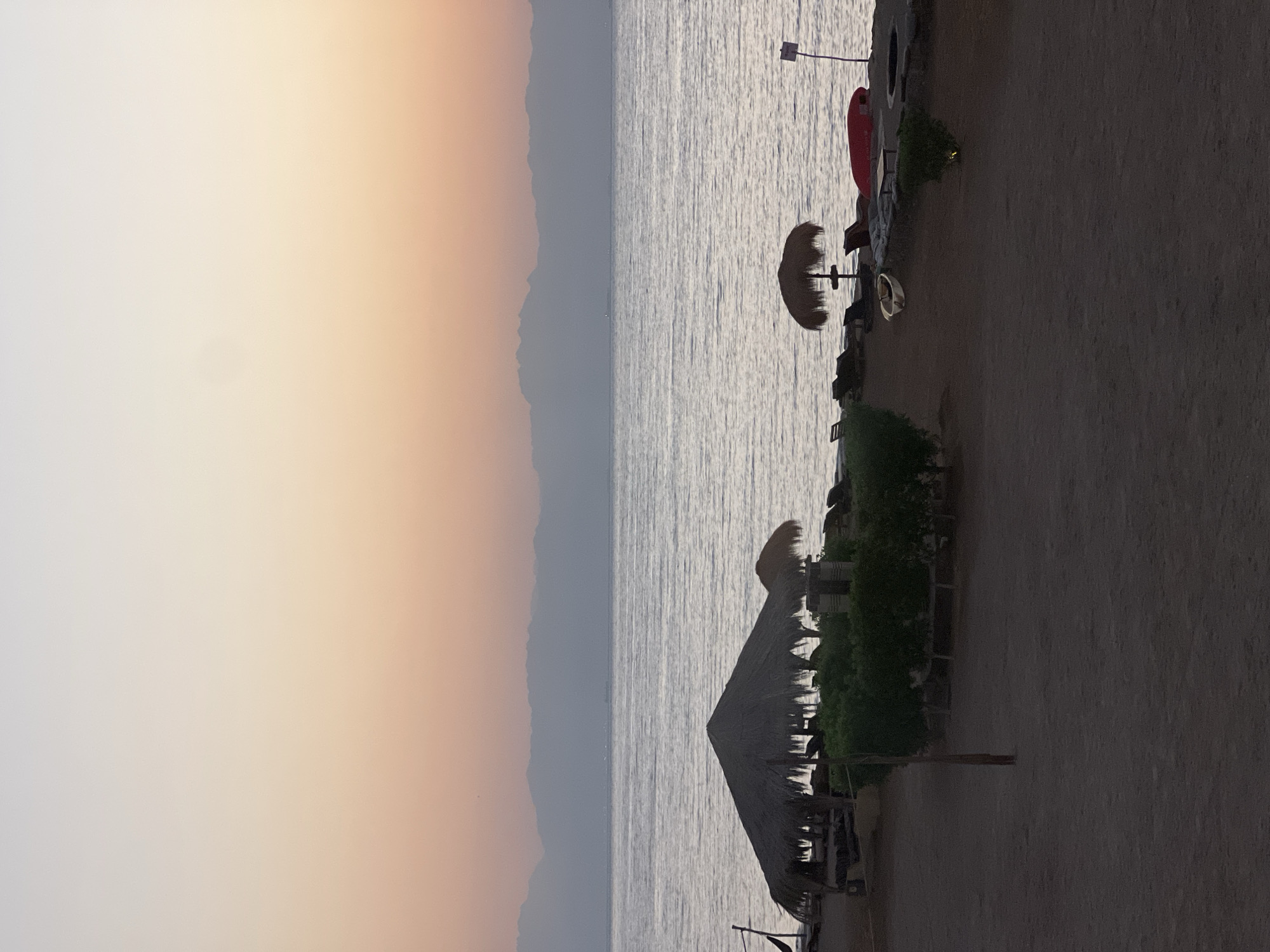 The image captures a serene beachfront scene in Nuweiba, Egypt, likely during sunrise or sunset, given the soft pastel hues of the sky. The foreground consists of sandy ground with sparse vegetation, including neatly trimmed bushes. Several thatched-roof umbrellas are scattered across the beach, providing shade for loungers and small tables. One prominent umbrella structure is larger and more elaborate, with a hammock visible to its left. A small, white boat rests on the sand near the water's edge, and a red kayak is positioned further down the beach. The calm sea stretches out to the horizon, where a faint silhouette of distant mountains can be observed. The overall atmosphere is tranquil and idyllic, suggesting a peaceful retreat.