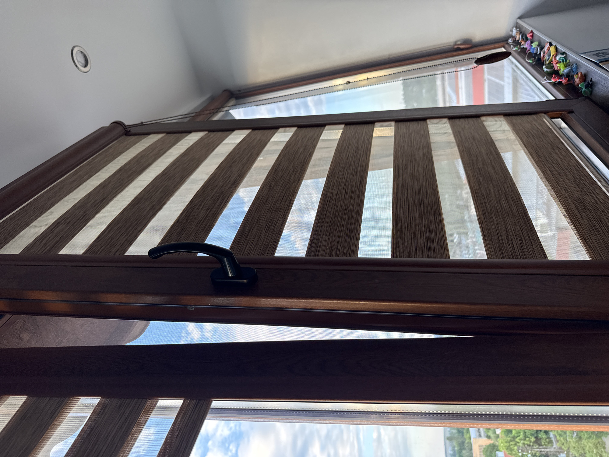 The image shows an open window with striped roller blinds in a room in Lovech, Bulgaria. The window frame is dark brown and the handle is black. The blinds alternate between brown and off-white stripes. Outside the window, the sky is visible with some clouds and a glimpse of greenery, possibly trees. On the top right corner of the window frame, there's a collection of small, colorful figurines. The ceiling is white with a recessed light fixture.
