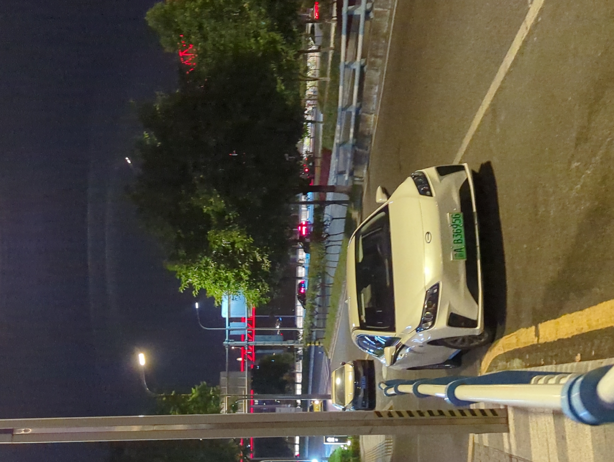 The image depicts a nighttime scene on a city street, likely in Jiangbei City Street, China, given the license plate format. In the foreground, a white electric vehicle with a green license plate reading "A B36956" is parked on the right side of the road. A portion of its front bumper, headlights, and hood are visible. A blue and white striped railing runs along the left side of the foreground, separating the sidewalk from the road. Further down the road, another car, appearing to be a yellow sedan, is in motion. Trees line the street, and in the background, there are streetlights and illuminated structures, including some with red accents. The sky is dark, indicating nighttime. The road surface appears wet in places, suggesting recent rain. The scene is predominantly illuminated by artificial lights from the street and vehicles.