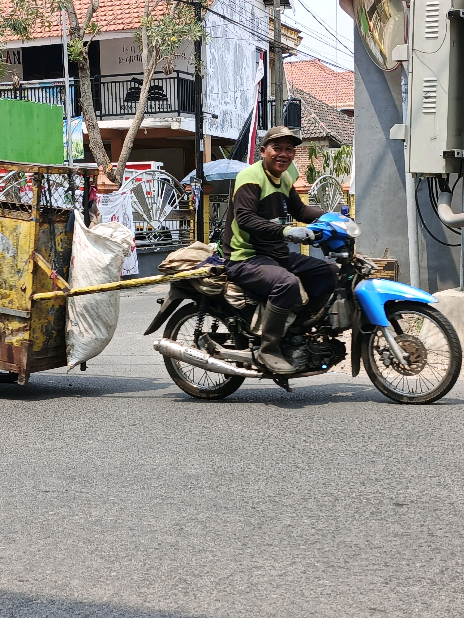 A man is riding a blue motorcycle with a trailer attached, on a street in Ketegan, Indonesia. The man is wearing a cap, a dark long-sleeved shirt with a green neckline, dark pants, and black boots. He is smiling and looking at the camera. The trailer is yellow and appears to be carrying bags of materials, one of which is a large white sack. In the background, there are buildings with red tile roofs, trees, and power lines. The scene is captured during the daytime, with the sun casting shadows on the road.