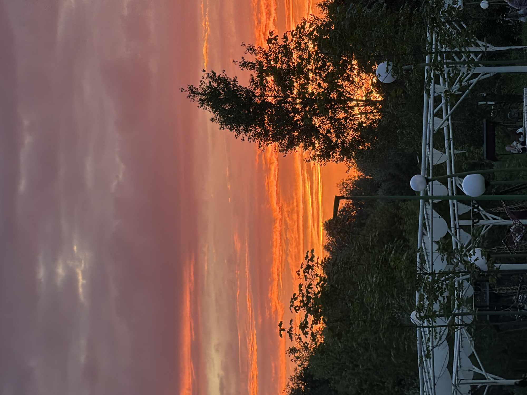 The image depicts a vibrant sunset over Kazan, Russia. The sky is filled with streaks of orange, pink, and purple clouds, indicating the sun is low on the horizon. Silhouetted trees and foliage frame the scene, with a white gazebo-like structure visible on the right side of the image, featuring round, white light fixtures. The overall mood is serene and picturesque, typical of an evening outdoors. No people are clearly visible, and there is no visible text in the image. The orientation of the image is portrait, but the scene itself is captured horizontally.