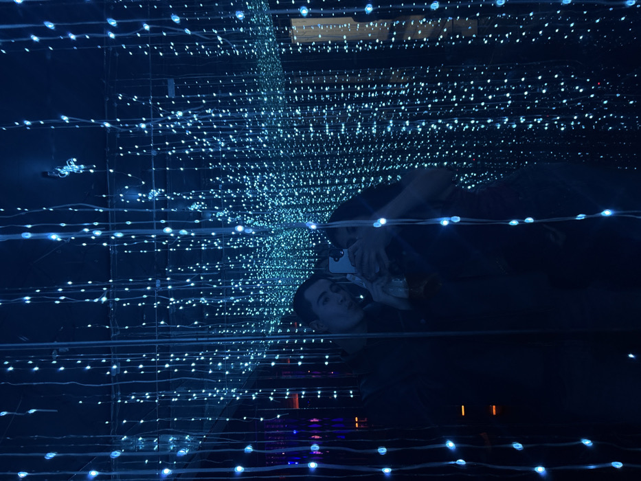 The image displays a reflection of two individuals captured within an indoor environment characterized by extensive blue LED string lights. Numerous vertical strands of small, glowing blue lights descend from the ceiling, creating a dense, immersive, and ethereal backdrop.
The subject on the left of the reflection is a male, appearing to be a young adult, with short dark hair. He looks towards the camera with his lips slightly puckered. He holds a clear glass bottle containing an amber liquid in his left hand. The subject on the right, partially obscured by a smartphone, is a female, also appearing to be a young adult. She is holding a light-colored smartphone with both hands, positioned to take a photograph, and her arm is around the male individual. Both individuals are wearing dark clothing. The reflective surface creates a sense of depth and amplifies the visual effect of the blue lights.
The environment appears to be a club, bar, or a specialized light installation designed for ambient display. The specific location within San Bernardino Tlaxcalancingo, Mexico, cannot be identified from the visual evidence provided.
