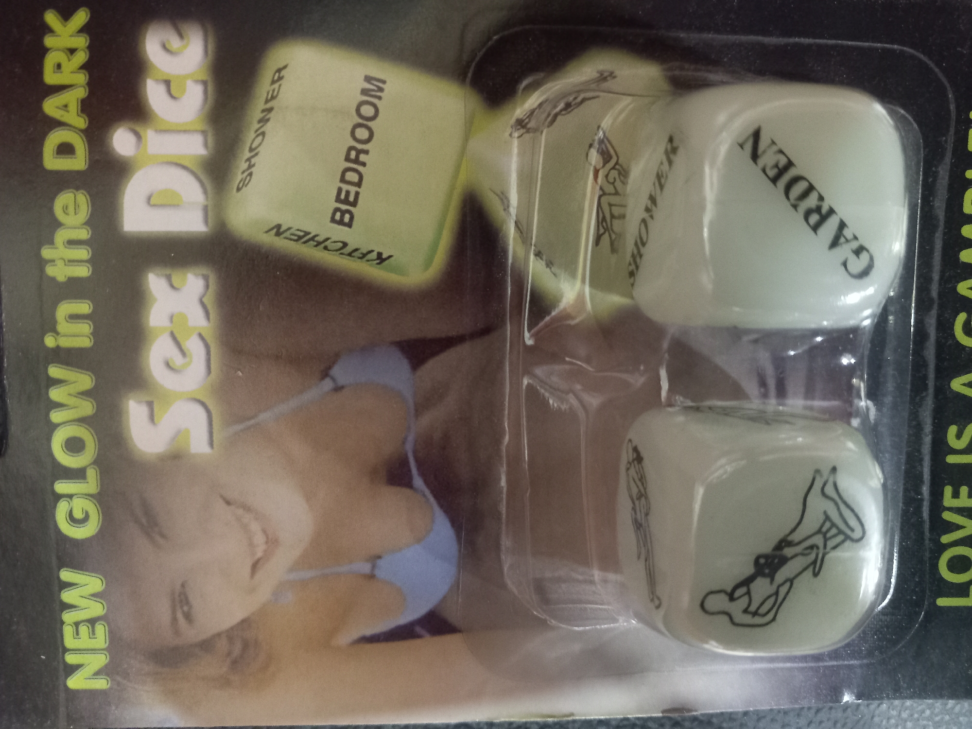 The image is a close-up shot of a product package for "NEW GLOW in the DARK Sex Dice." The packaging is made of clear plastic, and the background is dark. In the foreground, there are two dice visible. The left die is white and has a black illustration of two figures in a sexual pose. The right die is also white and has the word "GARDEN" printed on one face. Behind the dice, there is a larger, translucent green die with text on its visible faces: "KITCHEN," "BEDROOM," and "SHOWER." A portion of the upper torso and face of a smiling woman, wearing a blue bikini top, is visible in the upper portion of the image, behind the dice. The bottom of the packaging displays text that is partially obscured but appears to read "LOVE IS A GAMBLE." The overall lighting suggests an indoor setting, and the product appears to be new and in its original packaging.