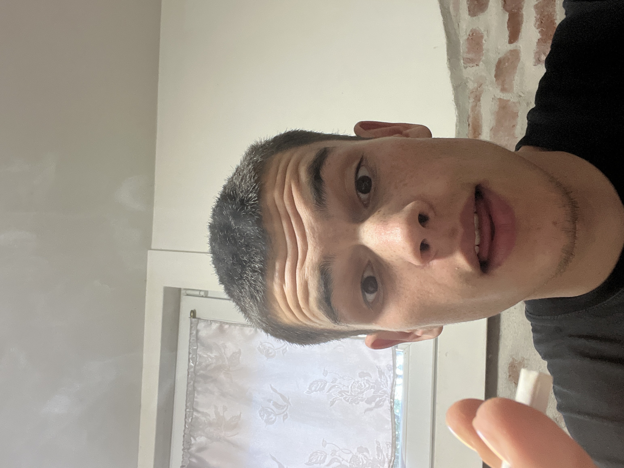 The image shows a selfie of a person with short, dark hair and light skin. The person has a visible brow line with multiple horizontal creases. They are wearing a dark-colored shirt. The background includes a white wall, a brick wall, and a window with a white curtain. The person's fingers are visible in the lower right corner, holding a small, white object. The location is not identifiable as Plovdiv, Bulgaria, based on the visible elements in the image.