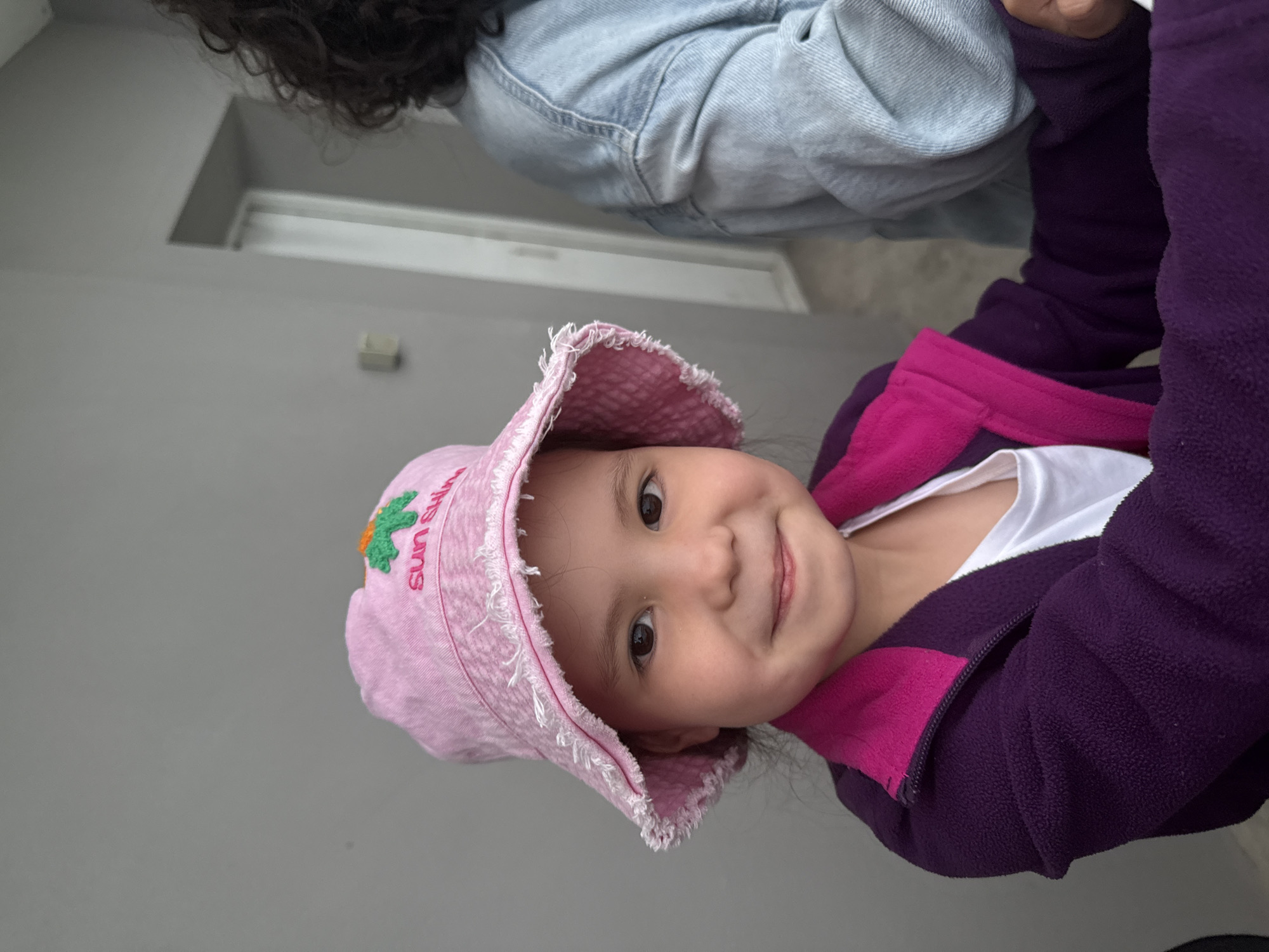 The image features a young girl, appearing to be between 4 and 7 years old, captured from the mid-chest up, with a slight smile and direct gaze towards the viewer. She is wearing a light pink bucket hat with a distressed, frayed brim. The hat displays green embroidered palm tree imagery and the text "sun shin" (likely "sun shine") on its front. Below the hat, she wears a dark purple fleece jacket with a vibrant pink inner collar, over a white t-shirt. To the girl's right, the shoulder and upper arm of another individual are partially visible, characterized by dark, curly hair and a light blue denim jacket. The background is a plain, light gray wall. On the right side of the frame, a segment of a white door frame or archway is visible, along with a small, light-colored rectangular fixture on the wall. The scene is consistent with an indoor or sheltered outdoor setting in the city of Perm, Russia.