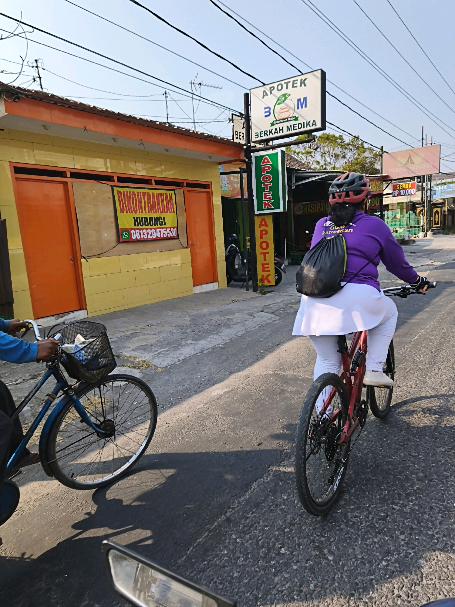 In Surakarta, Indonesia, on a sunny day, two individuals are cycling on a paved road. The foreground shows a person riding a blue bicycle with a basket filled with items. In the mid-ground, a person wearing a purple long-sleeved shirt, white pants, a white skirt, and a red helmet is riding a red bicycle. They are carrying a black backpack. To the left, a yellow building with orange doors has a sign that reads "DIKONTRAKKAN HUBUNGI 081329475530". Above the building and road, electrical wires and poles are visible against a clear blue sky. To the right of the yellow building, there are signs for "APOTEK 3M BERKAH MEDIKA" and other businesses. The road surface is asphalt with some cracks and debris. Shadows are cast on the road by the bicycles and buildings.