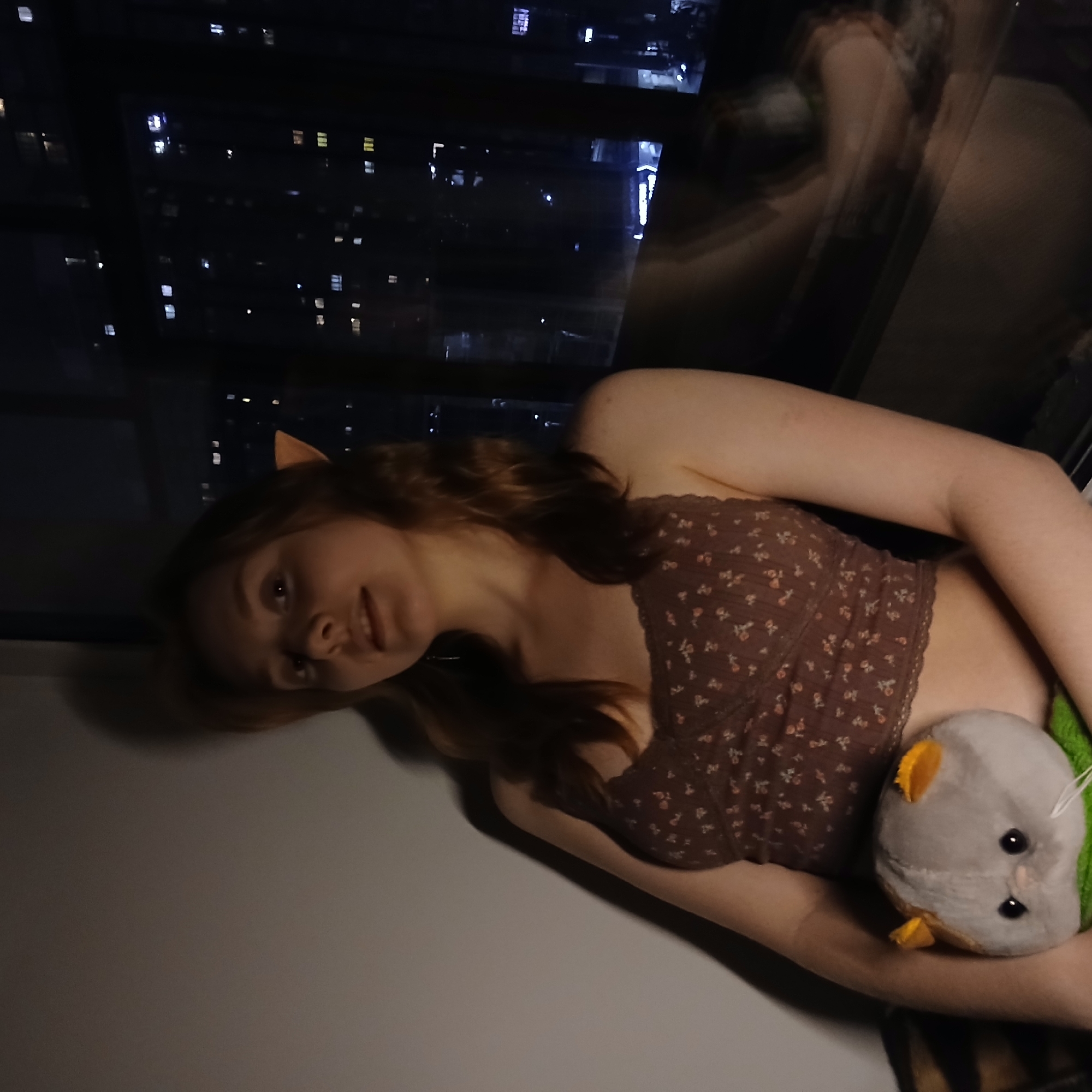 A young person with reddish-brown hair and light brown cat ears is lying down indoors, positioned alongside a large window overlooking a city at night. They are wearing a brown floral top with small white and pink patterns and lace trim. In their arms, they are holding a gray plush toy with yellow ears and feet, and black button eyes. The person looks towards the camera with a subtle, relaxed expression.
Through the window, the dark night sky contrasts with the numerous illuminated windows and structures of a cityscape, characteristic of Yekaterinburg, Russia. Faint reflections of the room and the person herself are visible in the window glass. The lighting in the room is dim, suggesting an intimate nighttime setting. No visible text is present.