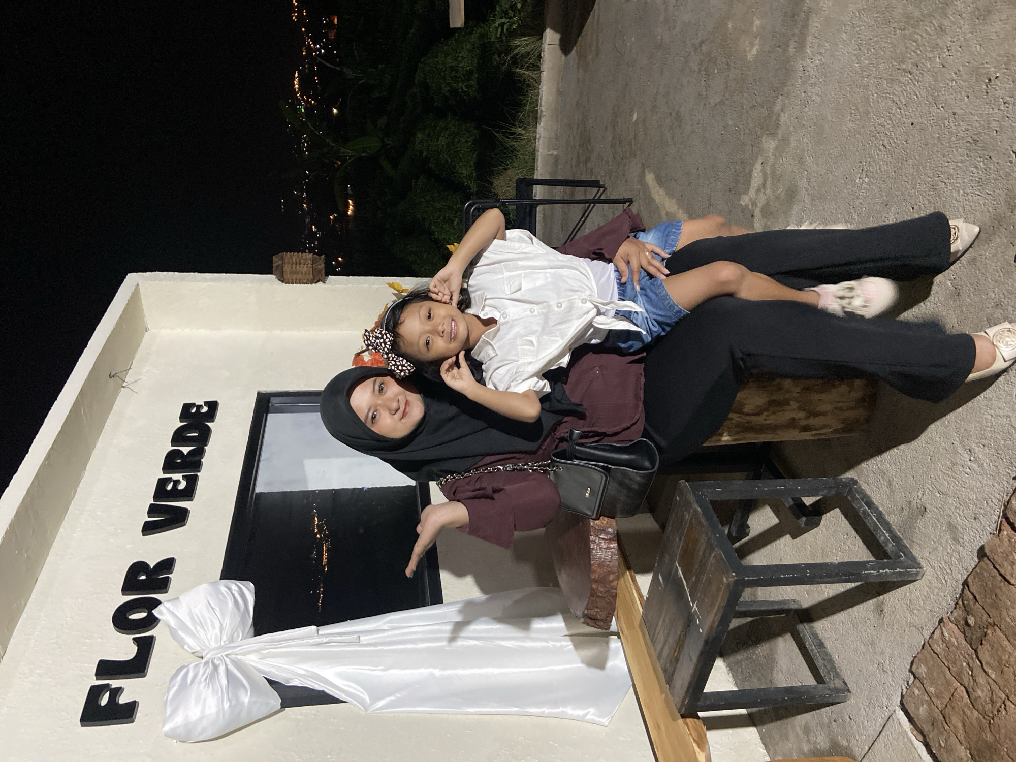 An adult female wearing a black hijab and a dark red top is seated on a log, holding a young girl on her lap. The girl is wearing a white shirt and denim shorts. Both are smiling at the camera. Behind them, a building has "FLOR VERDE" written in black letters on its exterior. A large white bow adorns the building near a window. The setting appears to be an outdoor seating area at night, with city lights visible in the background. The location is identified as Kesiman, Indonesia.