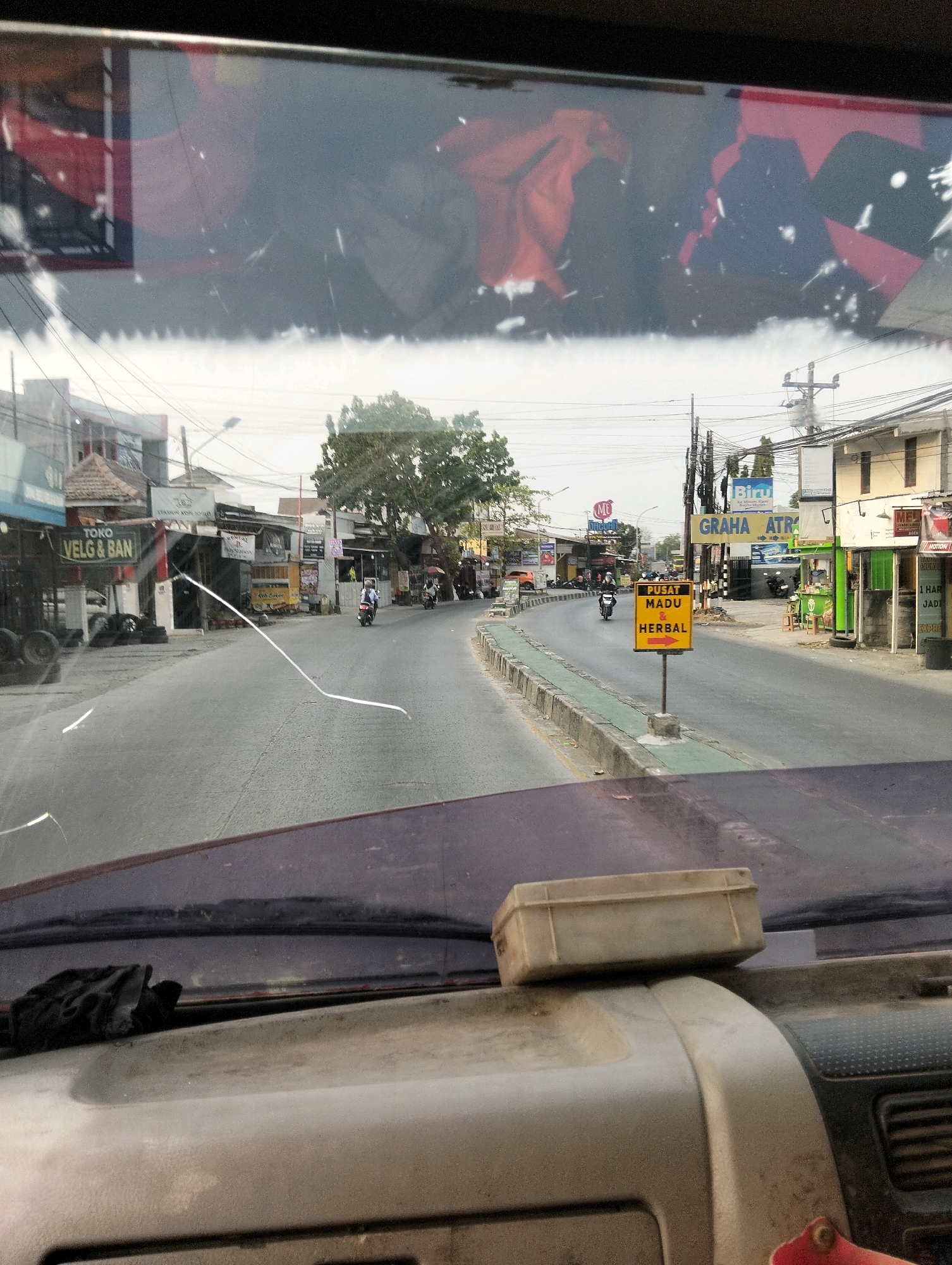 The image is a dashboard view from inside a vehicle looking out through the windshield at a street scene in Semarang, Indonesia. The windshield is dirty and has several cracks, including a prominent white line across the lower half. On the dashboard, a small beige rectangular box is placed on top of a grey surface, and a black cloth is resting on the left side.
The street ahead is paved and appears to be a moderately busy thoroughfare. On the left side of the road, there are several storefronts, including one with a sign reading "TOKO VELG & BAN" (Tire & Rim Shop). Tires are stacked outside this shop. Further down the street, more shops and buildings line the road, some with signage in Indonesian. A tree with green foliage is visible in the middle distance.
On the right side of the road, there are more buildings and shops. A prominent yellow sign with red text and a red arrow pointing to the right reads "PUSAT MADU & HERBAL" (Honey & Herbal Center). Other signs visible include "GRAHA ATR" and "Biru." Utility poles with numerous wires are present along the street.
Several motorcycles are on the road. One motorcycle with a rider wearing a helmet is in the foreground on the left lane, and another motorcycle is further ahead on the right lane. The sky is overcast, and the overall lighting suggests it is daytime. The architecture of the buildings and the signage indicate an urban or semi-urban environment in Indonesia.