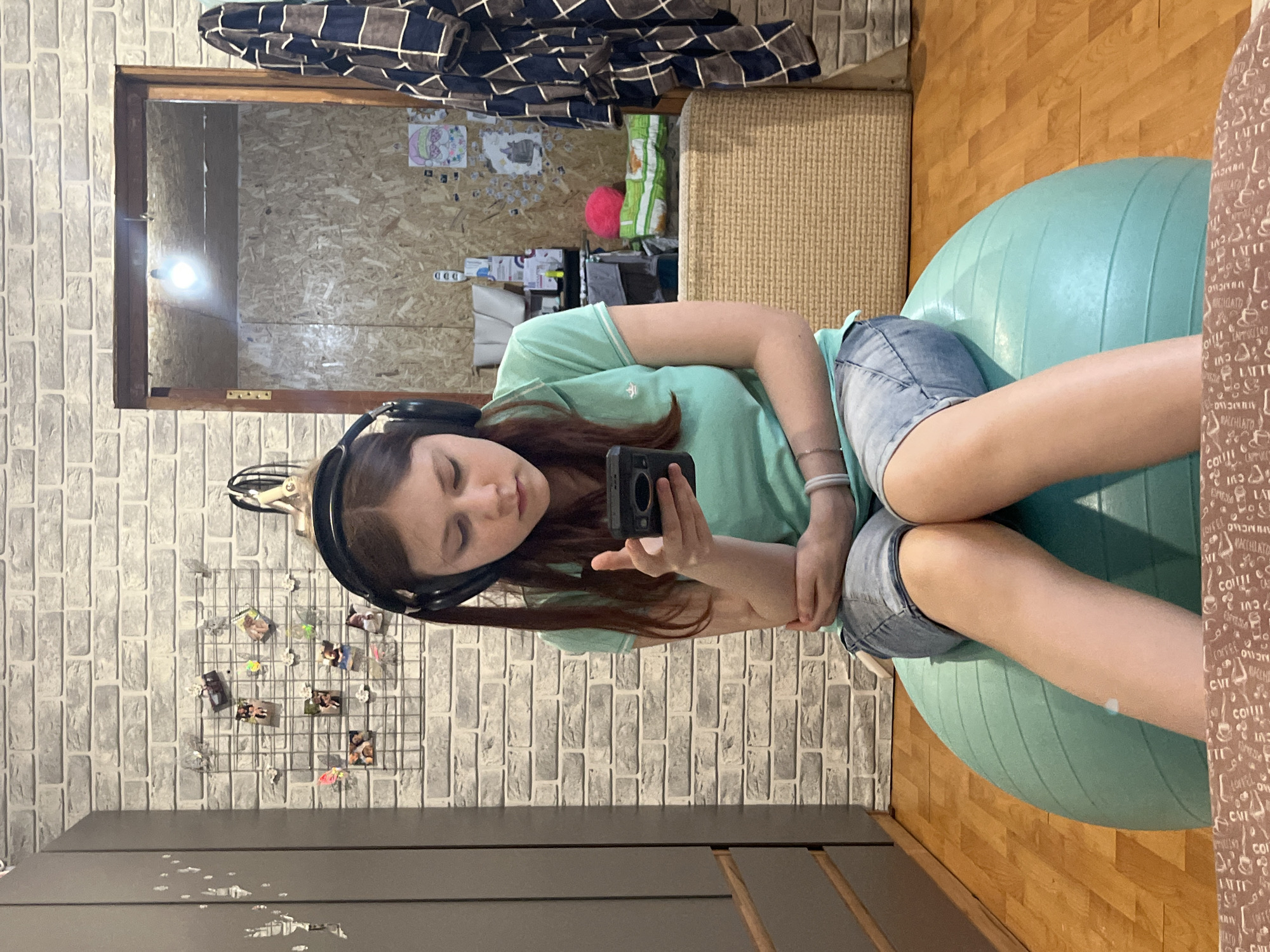 The image captures a young female indoors, likely in Vesyoloye, Russia, sitting on a large, light blue exercise ball. She wears a mint green t-shirt, denim shorts, and black over-ear headphones, looking down at a black smartphone or small camera held in her hands. Her expression appears calm and focused on the device.
The background features brick-patterned wallpaper. To the left, a metal grid adorns the wall, displaying several small photographs and mementos. A dark piece of furniture, possibly a dresser or cabinet, is partially visible at the bottom of the frame.
A prominent wooden-framed mirror in the upper left reflects another section of the room. This reflected area shows walls made of light-colored wood panels (possibly OSB or plywood) and a brightly glowing lightbulb. Drawings and pictures are taped to the reflected wall, and the letters "DDD" are visible below them. Also reflected are a light-colored woven object, perhaps part of a chair or basket, and a dark-patterned blanket or throw. The right side of the image reveals a wooden floor and a partial view of a brown-patterned fabric, possibly upholstery or wallpaper, adorned with various coffee-related words such as "COFFEE," "LATTE," "MACCHIATO," "CAPPUCCINO," "MILK," and "CUP." The scene suggests a casual and private setting, possibly a bedroom or living area.