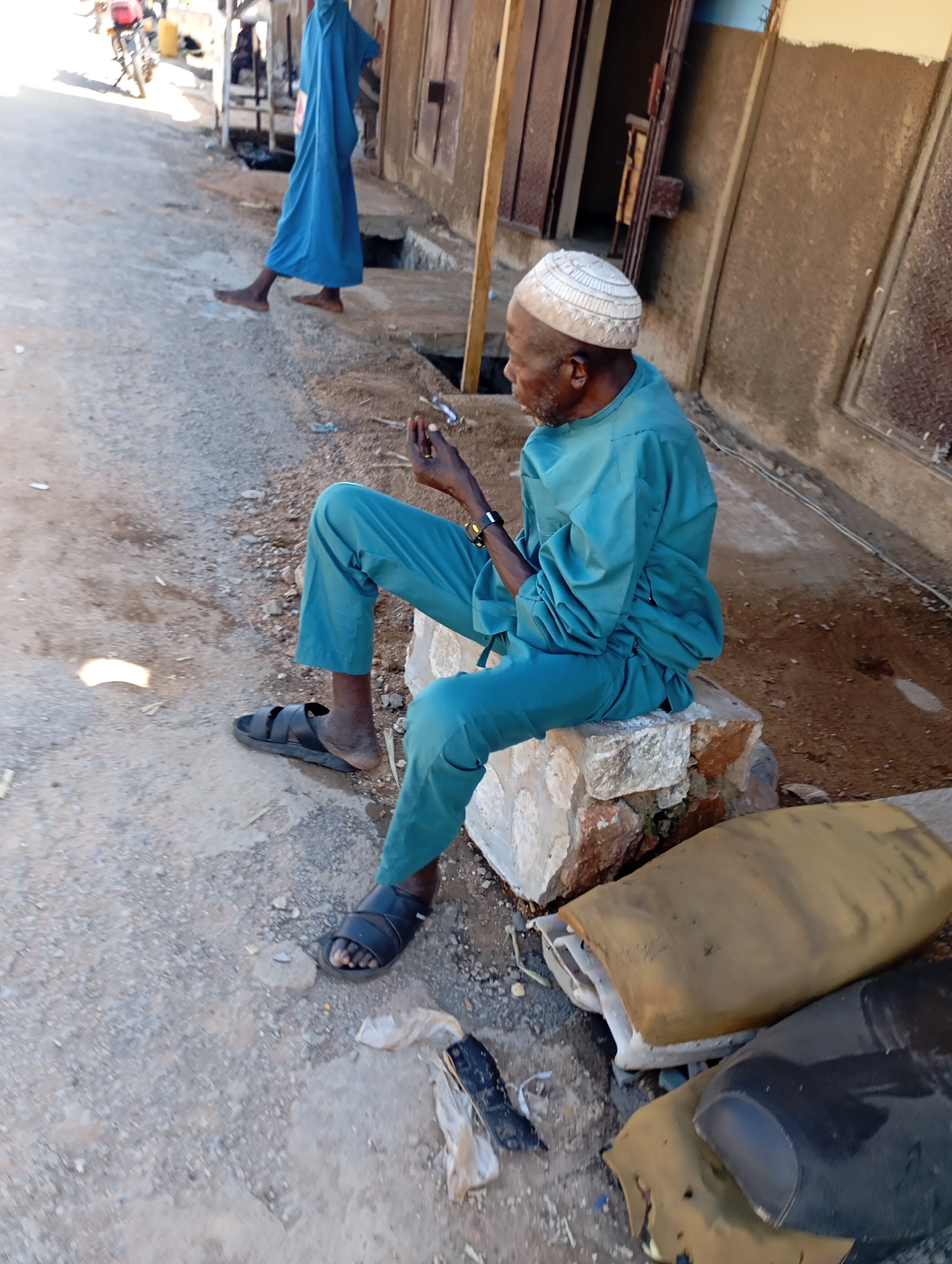 An elderly Black man wearing a teal garment and a white kufi hat sits on a rock in front of a building in Shendam, Nigeria. He is holding a small object, possibly a cigarette or needle, in his right hand and appears to be smoking or injecting himself. His left hand rests on his leg. He wears black sandals on both feet. The ground is paved with gravel and dirt. In the background, a person wearing a blue robe walks away from the camera. There are also several worn-out seat cushions scattered on the ground. The building in the background has a metal door and a brown facade. The scene appears to be an outdoor market or street setting.