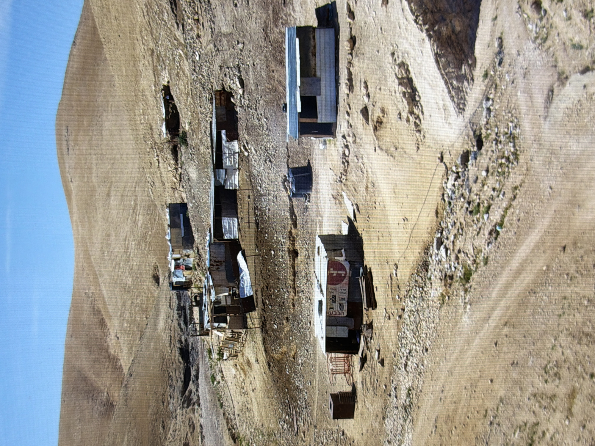 The image depicts a cluster of rudimentary dwellings on a dry, rocky hillside under a clear blue sky. The structures appear to be constructed from corrugated metal, wood, and possibly tarps, suggesting a makeshift or informal settlement. Some of the buildings have sloped metal roofs, and others are more basic, box-like structures. The terrain is arid and barren, with sparse vegetation visible in some areas. A dirt path winds through the scene, leading towards the cluster of dwellings. In the foreground, there are some scattered rocks and debris. In the middle ground, several buildings are visible, including one with a red and white banner. In the background, the hillside continues to slope upwards, with more structures scattered across it. The overall impression is one of a remote and undeveloped area. There is no indication of this scene being located in the city of Kyiv, Ukraine.