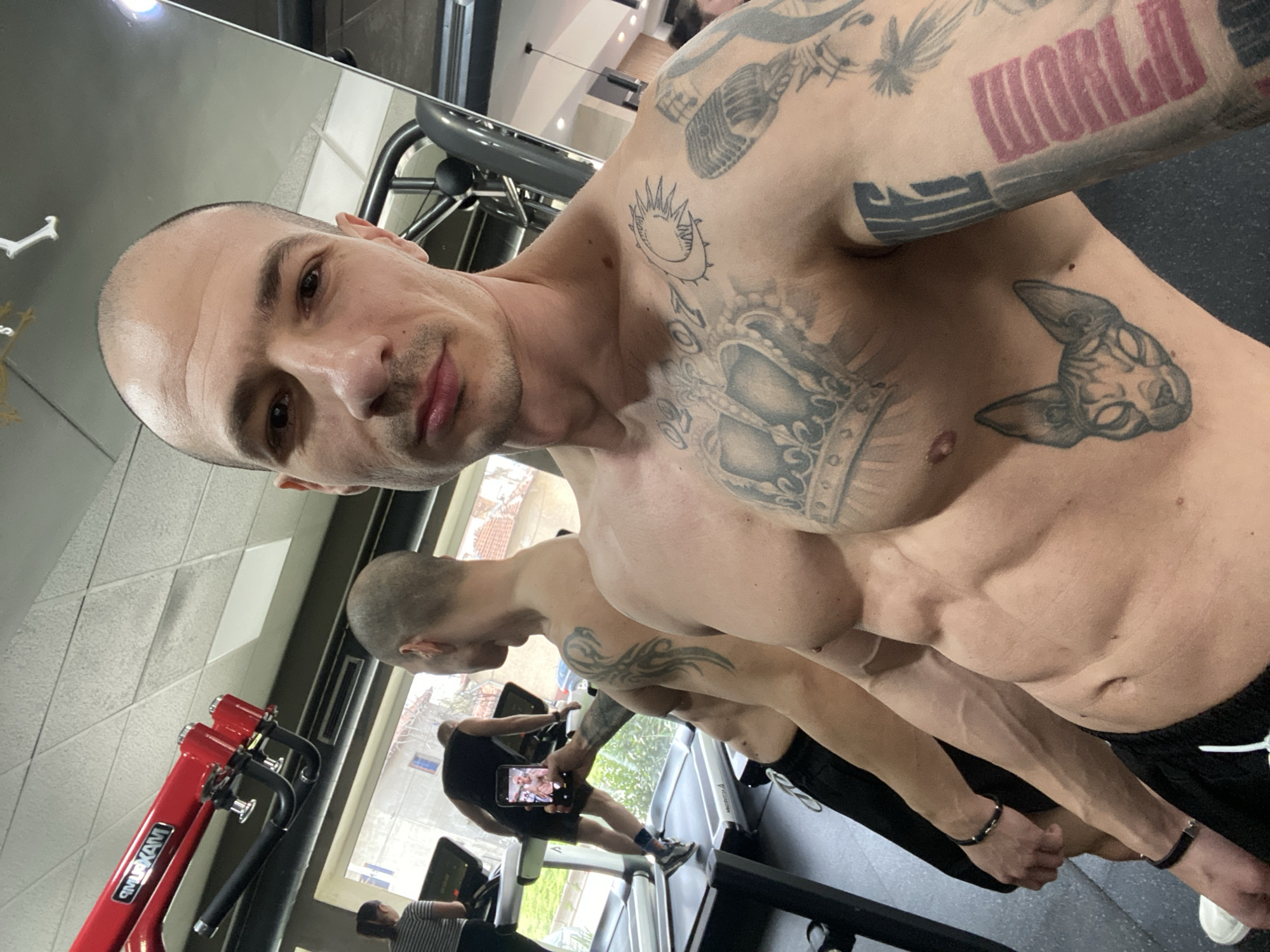 The image displays a shirtless, bald male subject taking a mirror selfie in a gymnasium. He has significant muscular definition, notably in the abdomen, and prominent vascularity on his arms. His attire consists of black shorts with white drawstrings and white sneakers. Black beaded bracelets are present on both wrists. The subject is extensively tattooed, featuring a large crown design centered on his chest, the numbers "02-07" near his right clavicle, a stylized sun and crescent moon on his upper right chest, and a Sphynx cat head on his left pectoral. His right arm displays a tattoo of a vintage microphone and abstract patterns, including red horizontal lines on the forearm. His left arm, visible in reflection, bears a tribal-style tattoo.
The background, primarily viewed through the mirror reflection, shows the interior of the gym and an exterior urban scene. A red "MAXPUMP" labeled exercise machine is visible overhead against a white tiled ceiling. Behind the main subject, a bald male, wearing a black tank top and black shorts, is walking on a treadmill. He holds a black smartphone horizontally, seemingly capturing an image or video. To his left, a female wearing a striped shirt is also on a treadmill. Beyond the treadmills, a large window provides a view of an exterior environment with residential buildings, one painted light blue, along with trees and what appears to be a street. The floor of the gym is dark. This scene is situated in a gymnasium in Pleven, Bulgaria.