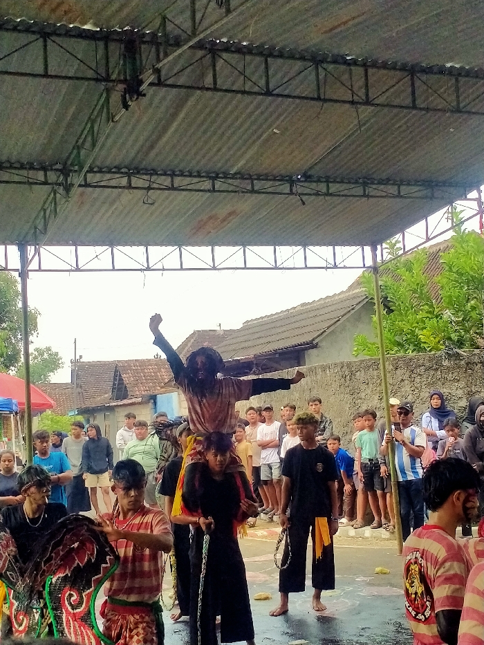 An outdoor cultural performance is captured under a corrugated metal roof structure in Jenggrik, Indonesia. Centrally, a figure with dark, matted hair and a dark face, wearing a light-colored, stained shirt, is carried on the shoulders of another individual. This elevated figure's arms are outstretched, forming a horizontal line. The carrier wears dark clothing with yellow and red fabric draped over their shoulders. To the left foreground, a performer in a black shirt, with dark smudges on their face, holds a large, ornate traditional mask exhibiting red, black, and white patterns. Another performer in a striped reddish-white shirt is partially visible to the right of the mask-holder. To the right midground, a barefoot individual in a black t-shirt and wide-leg dark pants with a yellow sash holds a chain. The ground beneath the performers is wet and marked with white patterns. In the mid-background, a crowd of spectators, primarily young adults and children, observes the event from behind a low concrete wall. Beyond the wall, several houses with tiled roofs and green vegetation are visible under an overcast sky. A spectator on the far right, wearing a light blue and white striped shirt, is holding a smartphone vertically. The performance area is covered by a corrugated metal roof supported by dark metal trusses.