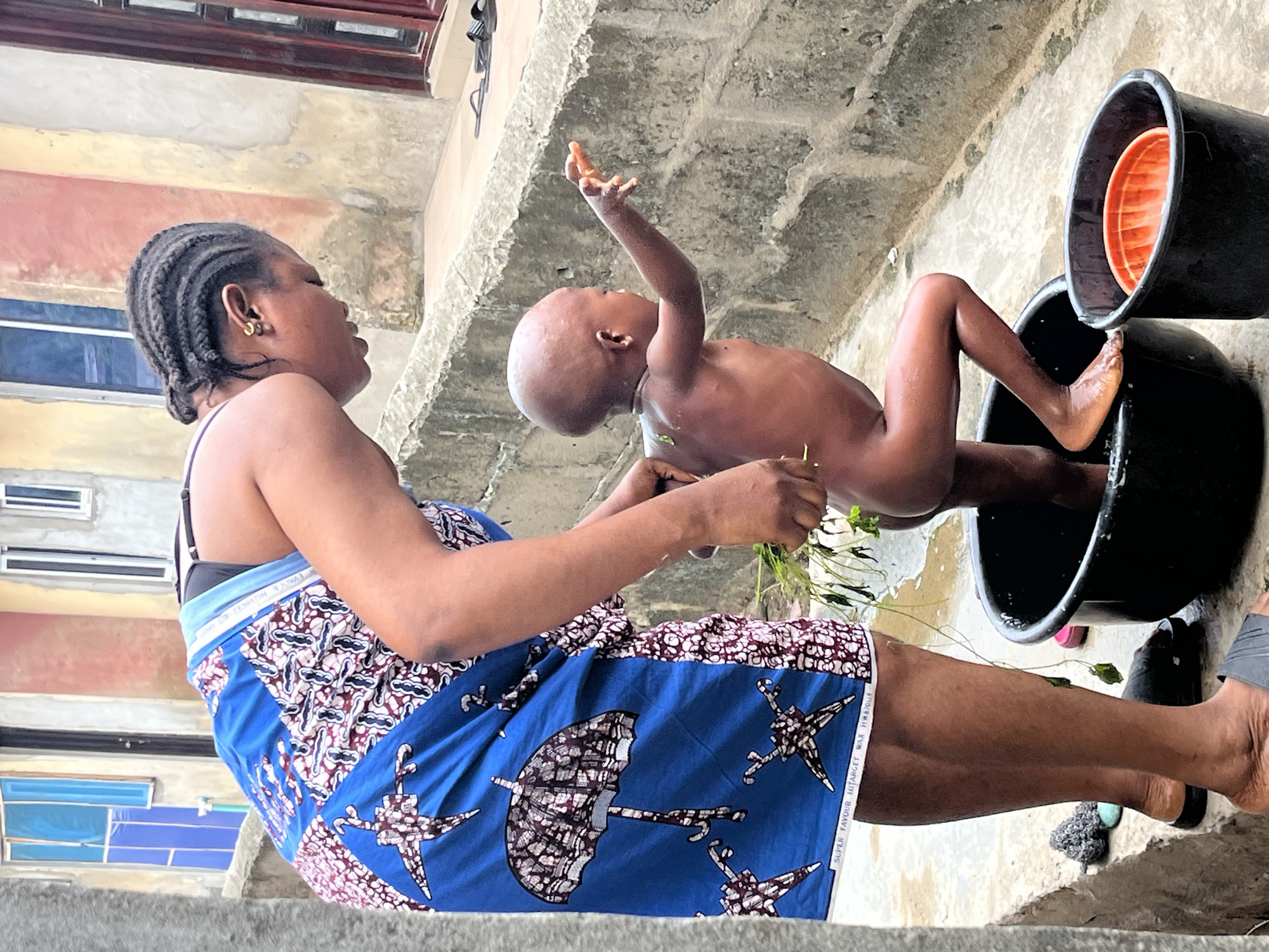 In Badagry, Nigeria, a woman bathes a baby outdoors using water from a black bucket. The baby sits upright in the bucket, legs extended. The woman, adorned in a blue and white patterned cloth, uses foliage to wash the baby. A second bucket, containing an orange container, is adjacent. The setting is a textured concrete courtyard next to a building.