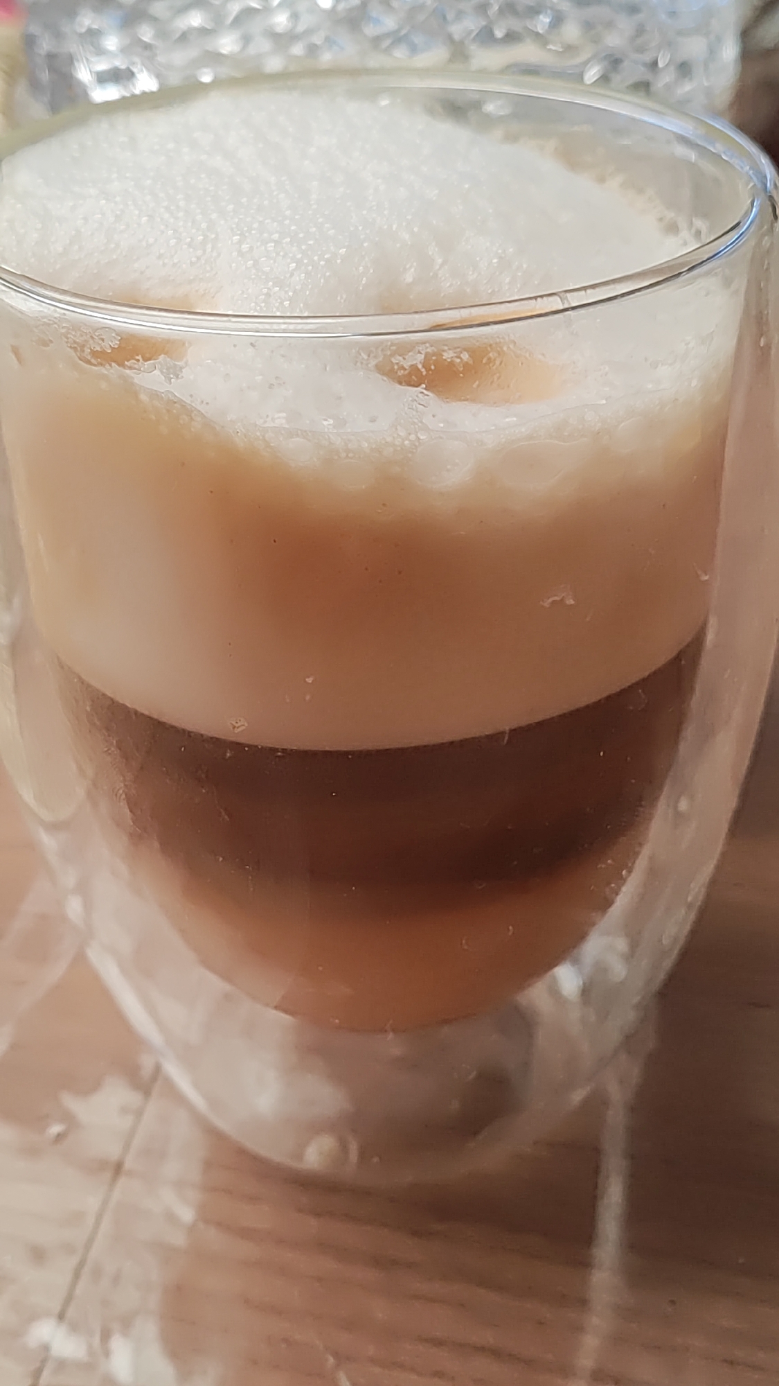 A close-up, vertical shot captures a layered beverage in a clear, double-walled glass. The drink displays distinct strata of colors: a dense, white foam crowns the top, followed by a lighter, creamy beige layer, then a darker, rich brown layer, and finally a lighter brown at the bottom. The glass itself has a rounded bottom and is set against a blurred background of a wooden surface. The overall impression is that of a well-prepared coffee or latte, presented for consumption. There are no discernible geographical markers that would indicate a specific location in Sochi, Russia.
