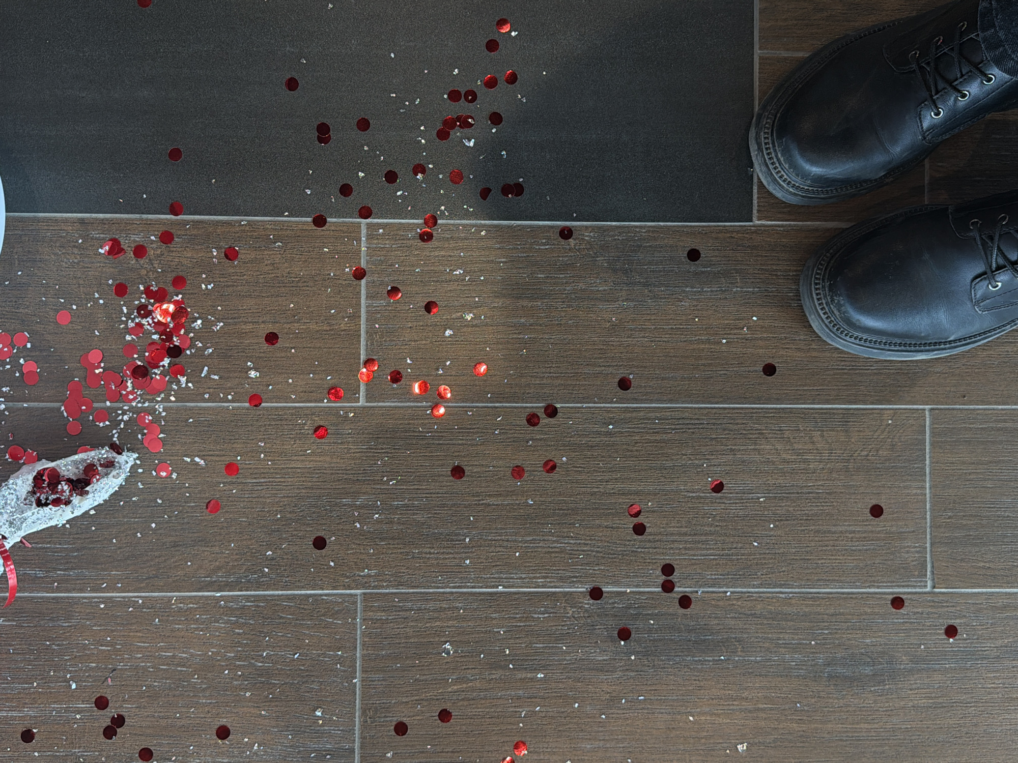 The image depicts a scene indoors. A portion of a broken champagne flute lies on its side on the left side of the frame, spilling its contents: small, round, red confetti and white, reflective glitter. The confetti and glitter are scattered across the floor. The floor is made of rectangular tiles arranged in a linear pattern. The floor material appears to be wood-look tile with shades of brown. To the right of the spilled confetti and glitter, a pair of black leather boots are visible. The floor tile pattern changes to a dark gray, solid surface near the top of the frame where the boots are located.
There is no information in the image indicating this location to be in Izhevsk, Russia.
