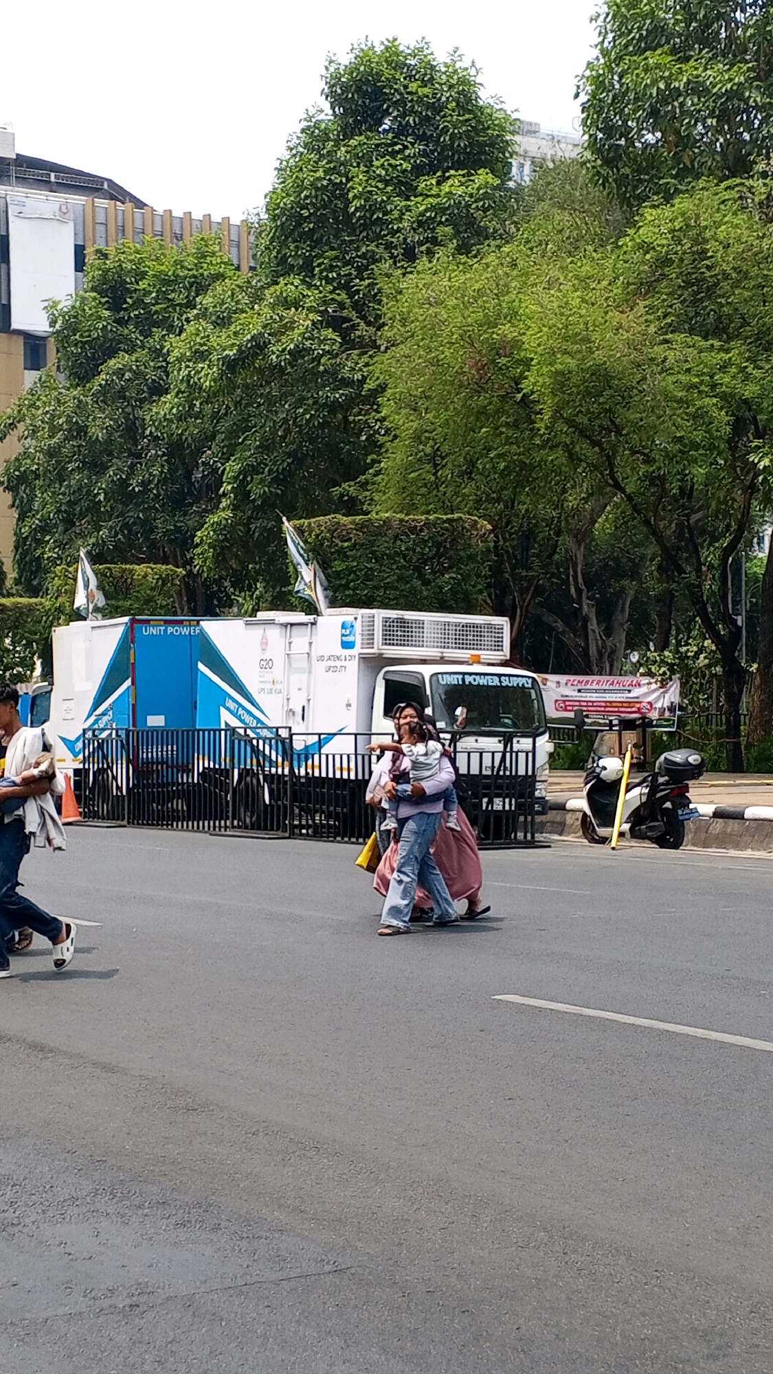 The image captures a street scene in Semarang, Indonesia, likely during daylight hours. A white truck with blue accents, labeled "UNIT POWER SUPPLY" and "G20", is parked on the road. Several people are crossing the street in front of the truck. On the right side of the road, a black scooter is parked. Lush green trees line the background, with a building visible behind them. The road surface shows signs of wear and tear.
