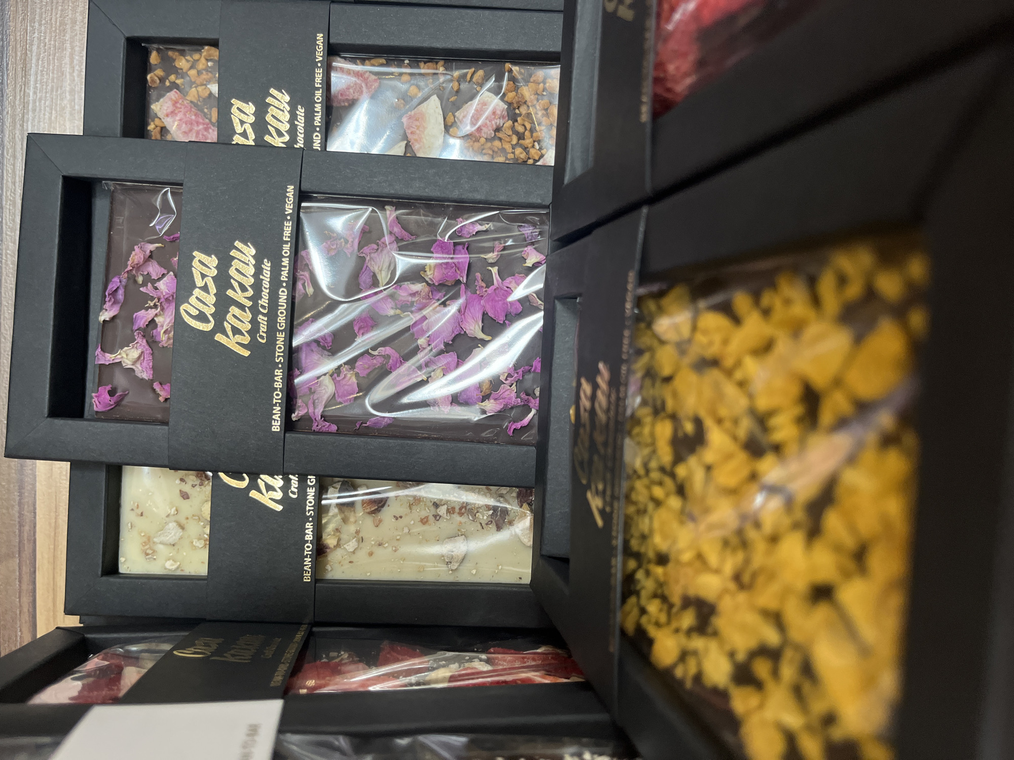 The image depicts a stack of chocolate bars from Casa Kakau, a craft chocolate maker. The bars are packaged in black boxes with clear windows, revealing various toppings on the chocolate. From top to bottom, the visible toppings include nuts and dried fruit, purple flower petals, white chocolate and nuts, and red fruit pieces. The text on the boxes indicates "Bean-to-Bar," "Stone Ground," "Palm Oil Free," and "Vegan." While the location isn't directly visible, Casa Kakau is based in Sofia, Bulgaria, suggesting the photo was taken in a retail setting or storage area there.