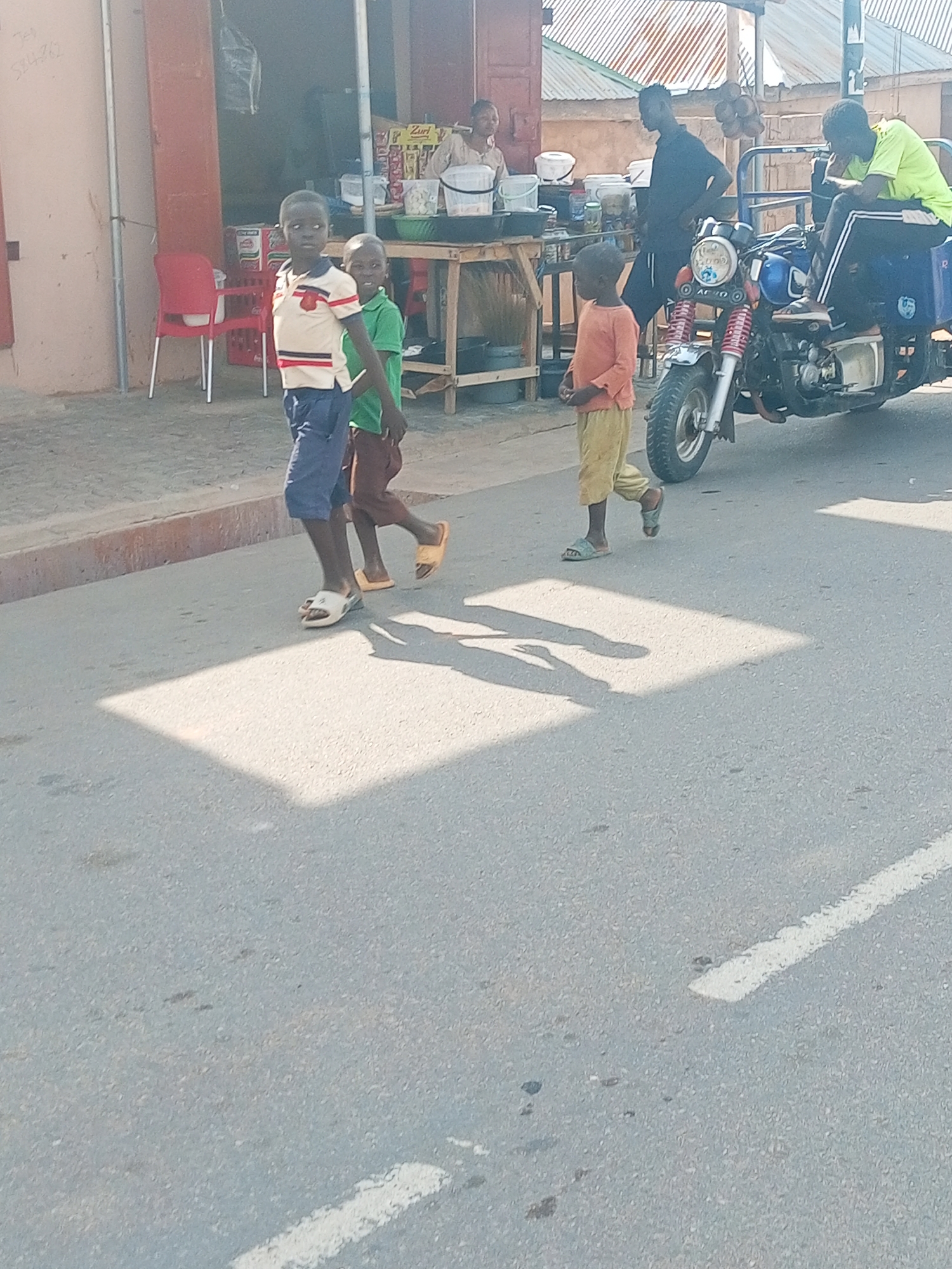 Three children are walking along the road in Gombe, Nigeria. One child wears a collared shirt, and the other two wear an orange and green T-shirt, respectively. In the background, there is a tricycle parked by a makeshift store with people. The background shows a partial view of buildings and a corrugated roof.