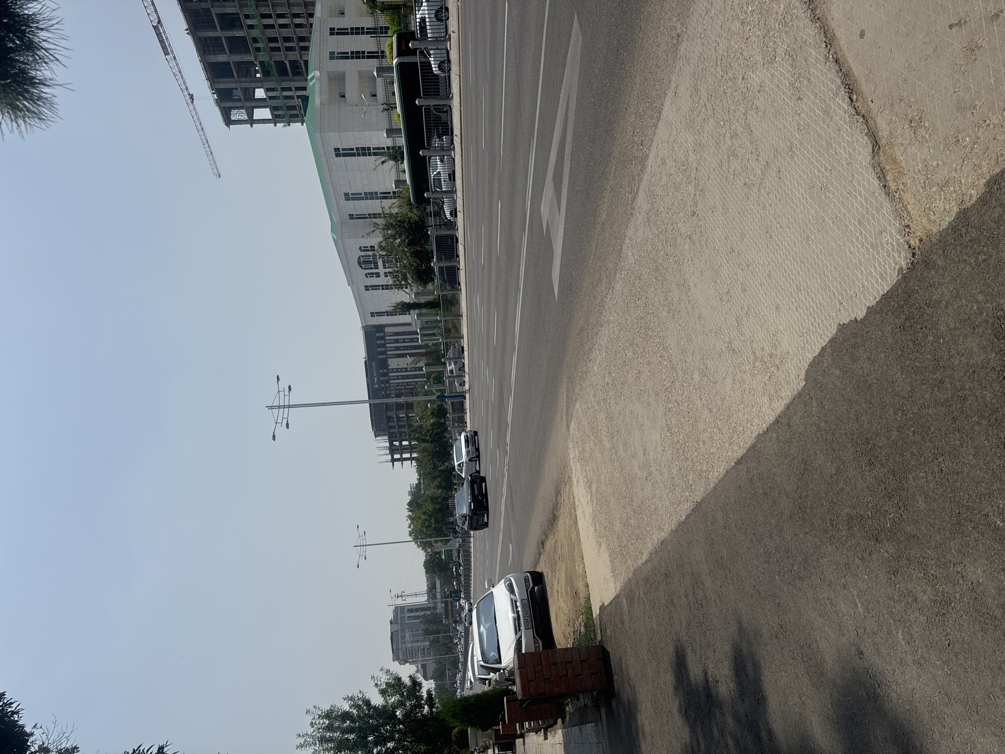 The image shows a street scene with a road, sidewalk, and buildings. The road has white lane markings and a white arrow painted on it. Several cars are parked along the side of the road. In the background, there are modern buildings, some of which appear to be under construction. A construction crane is visible against the sky. The sky is clear and hazy, suggesting it might be a warm day. The light indicates it is daytime. The perspective of the image is from a low angle, looking across the road.