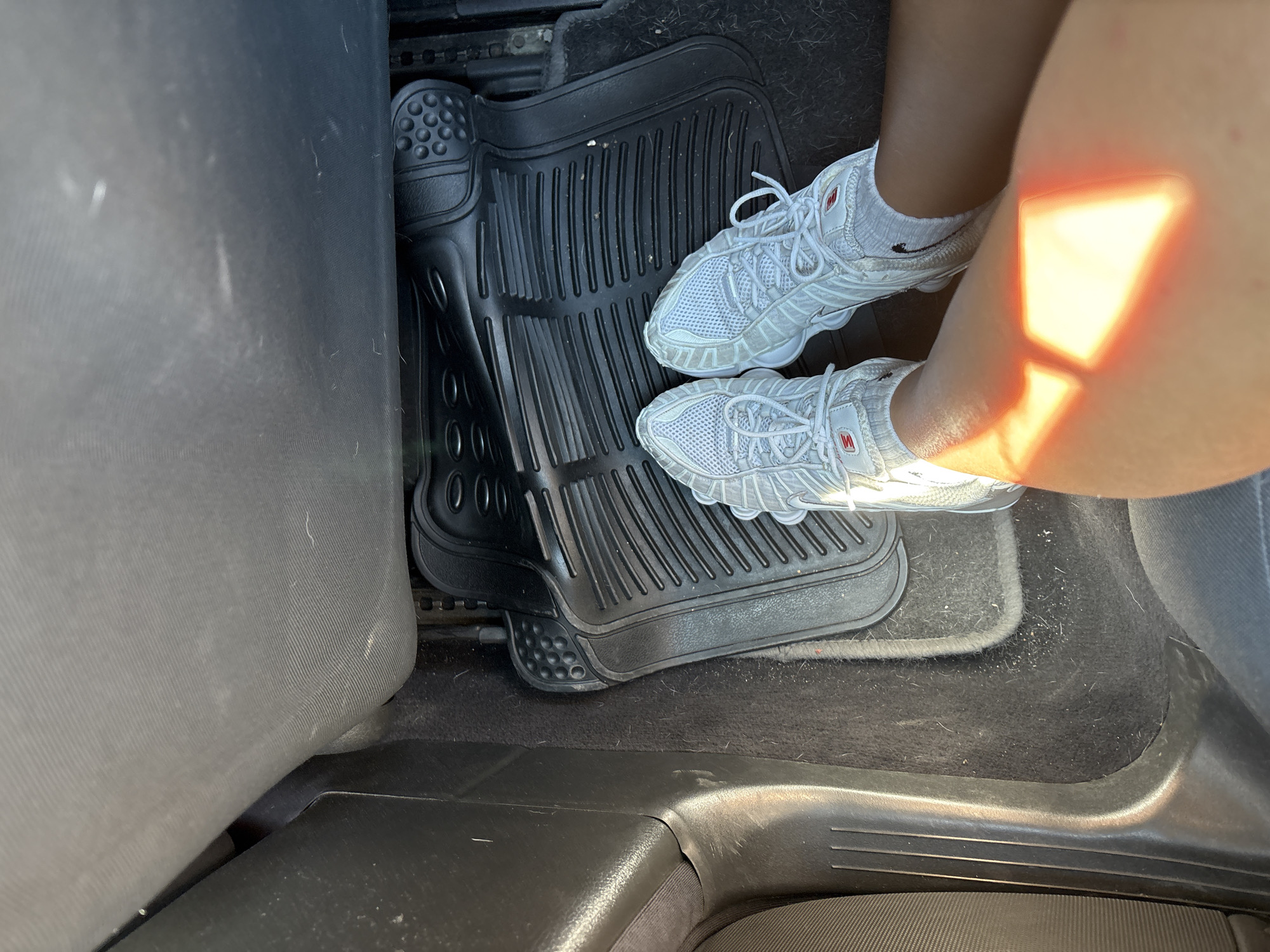 A pair of white sneakers and legs are visible inside a vehicle, positioned on a black rubber floor mat. The floor mat is ribbed and appears to be a custom fit for the car. The interior of the car is dimly lit, with sunlight casting a bright orange rectangular reflection on the visible leg. The background consists of the car's upholstery and plastic trim. The image is taken from a high-angle perspective, looking down at the feet and floor of the vehicle.
