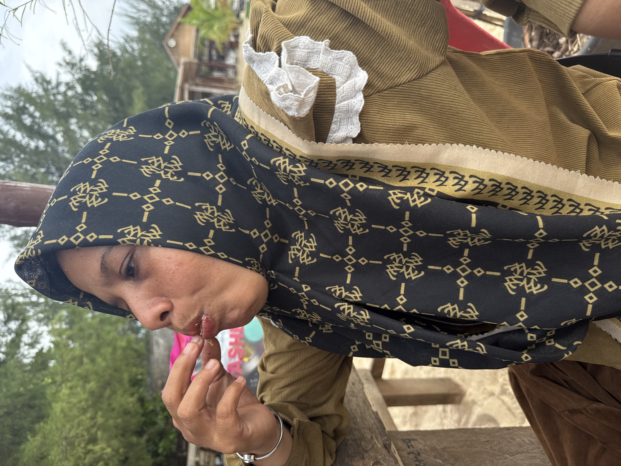 This is a medium shot photograph of a woman wearing a headscarf and a brown top. She is looking down and to her left, with her right hand raised to her mouth, as if eating or about to eat something. Her lips are slightly parted and appear to have some red lipstick on them. She is wearing a black headscarf with a gold geometric pattern. The top she is wearing is a textured brown fabric, and she has a white ruffled embellishment on the hood of her headscarf. A silver bracelet is visible on her wrist.
The background is slightly blurred, but appears to be an outdoor setting with trees and some wooden structures. The lighting suggests it is daytime, possibly overcast. The woman's expression is neutral. The scene is set outdoors, with the background suggesting a natural or rustic environment.