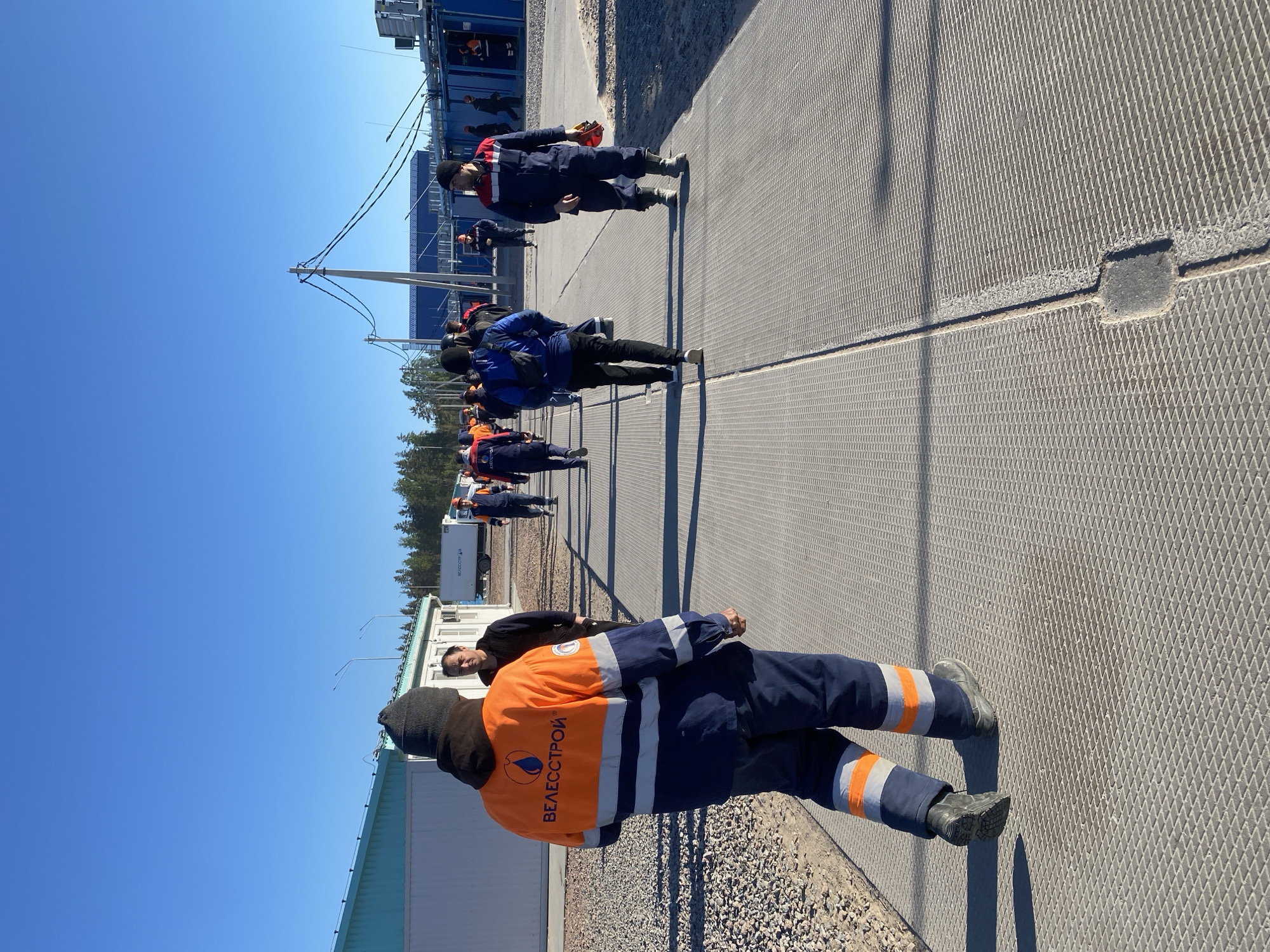 A group of people in blue and orange work suits are walking along a gray metal grate pathway. The people appear to be near a building, with a clear blue sky overhead. The location is Koskolovo, Russia, based on the text printed on the back of the nearest person.