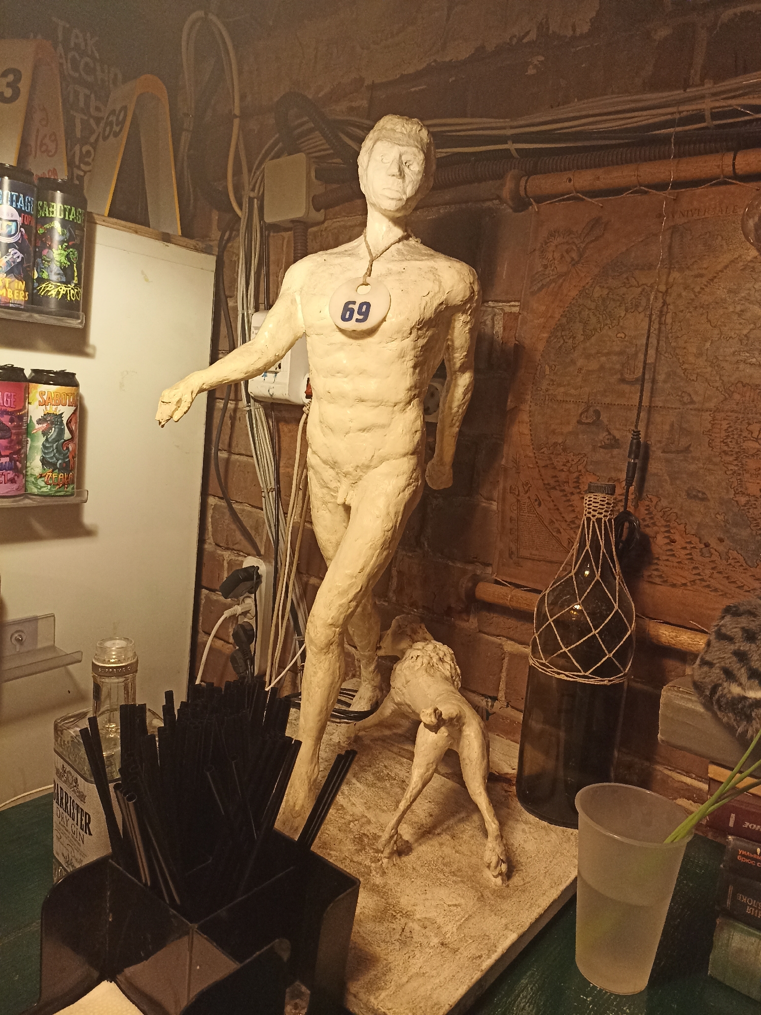 The image appears to be a cluttered interior space in Perm, Russia. The main subject is a nude male statue with a "69" tag around its neck. A clay dog figure sits near its feet. Behind the statue is a brick wall with exposed electrical cables. To the right of the statue is a dark bottle-lamp hanging in front of an old map. A clear plastic cup with green stems stands next to the lamp. In the foreground, there is a container filled with black straws next to a bottle labeled "BARRISTER DRY GIN". Cans of "SABOTAGE" energy drinks are visible on a shelf to the left of the statue. The overall scene is dimly lit and appears to be in a bar or pub.