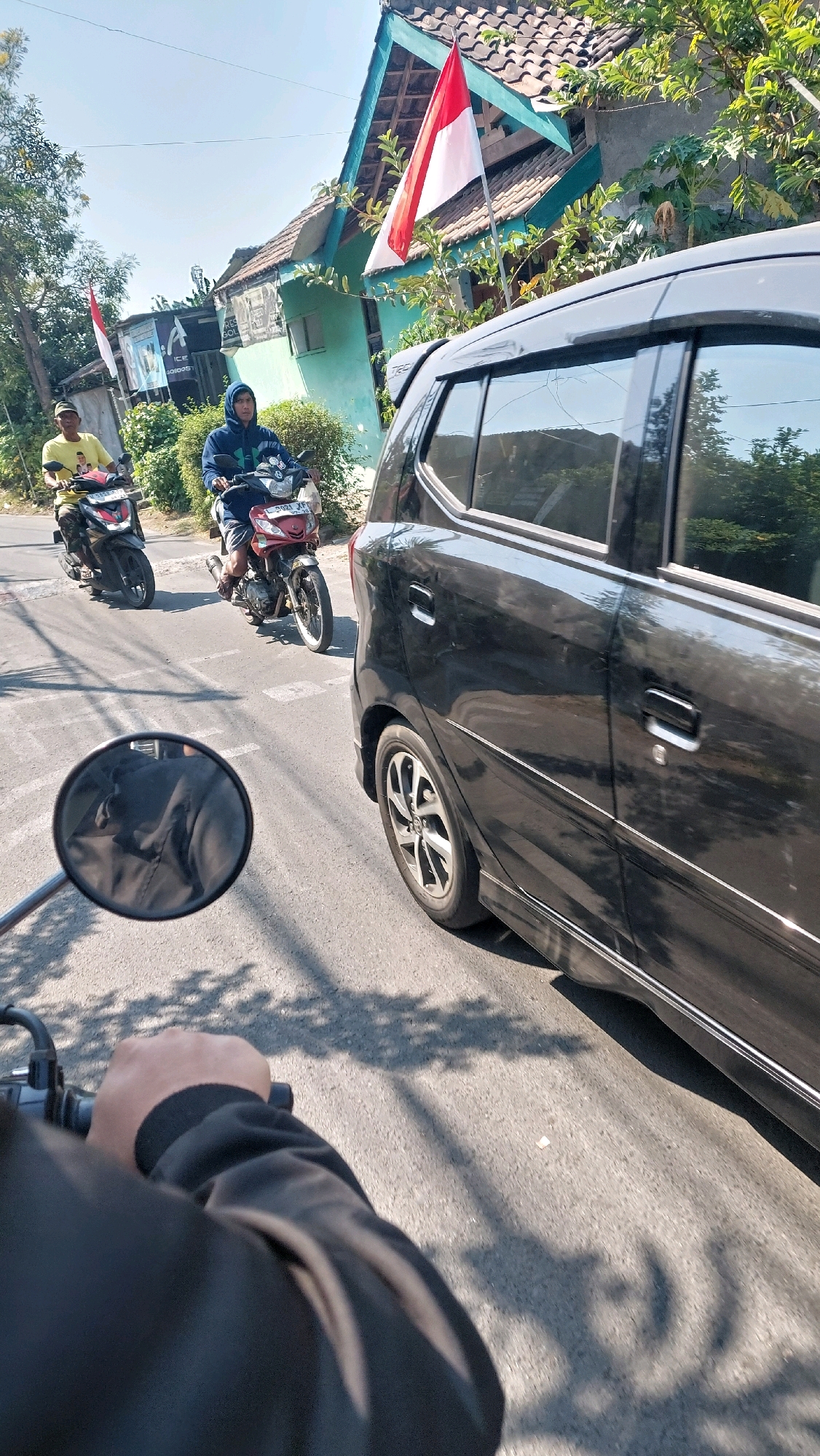 In Sidoarjo, Indonesia, on a sunny day, two motorcycles and a black car are traveling on a paved road. The first motorcycle, ridden by a man in a yellow t-shirt, is in the left lane. The second motorcycle, carrying a person in a blue hooded sweatshirt, is in the right lane. Behind them, a black car is also proceeding down the road. Trees line the street, and houses with red and white flags are visible in the background, suggesting a national celebration. The image is captured from the perspective of someone on another motorcycle, with their arm and a rearview mirror visible in the foreground.