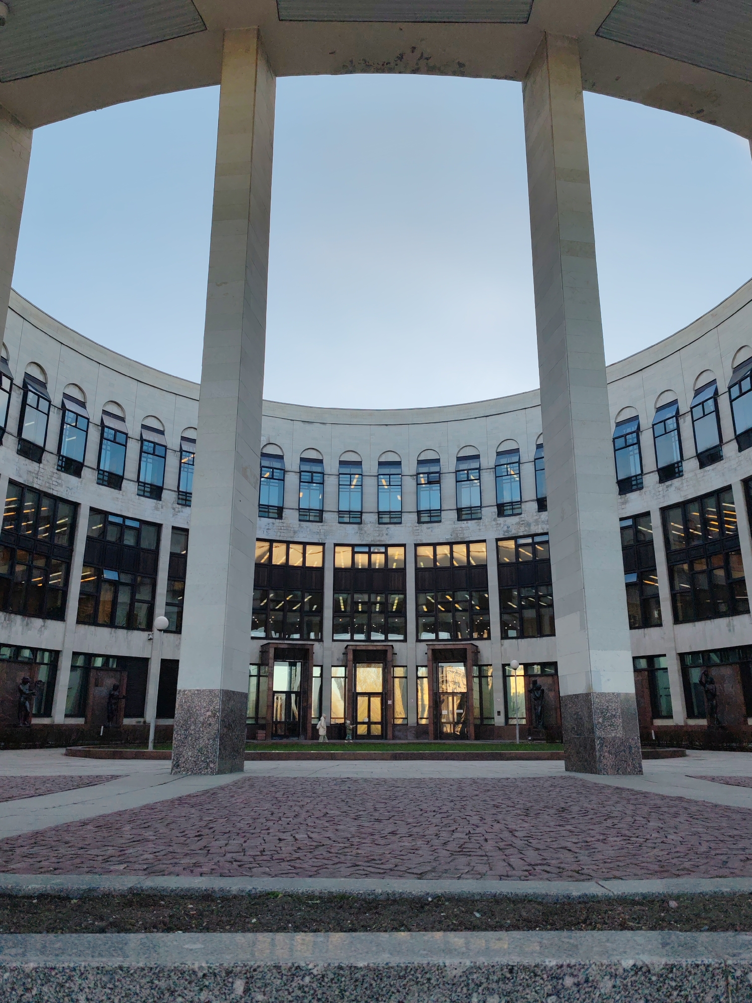 The image presents a low-angle, eye-level view of an architectural structure located in Saint Petersburg, Russia. The building features a curved, multi-story façade with arched windows and prominent support columns. The building's exterior is primarily composed of light-colored stone or concrete. Dark-framed windows reflect an orange glow, likely indicating the presence of interior lighting or a sunset. Below the windows on the ground level, there are doorways and statue niches on the ground level.
The foreground is a paved courtyard constructed of red paving stones. Two support columns are in the foreground, with the sky visible above the building. The sky is pale blue, suggesting either dawn or dusk. The overall impression is one of classical design and symmetrical construction.