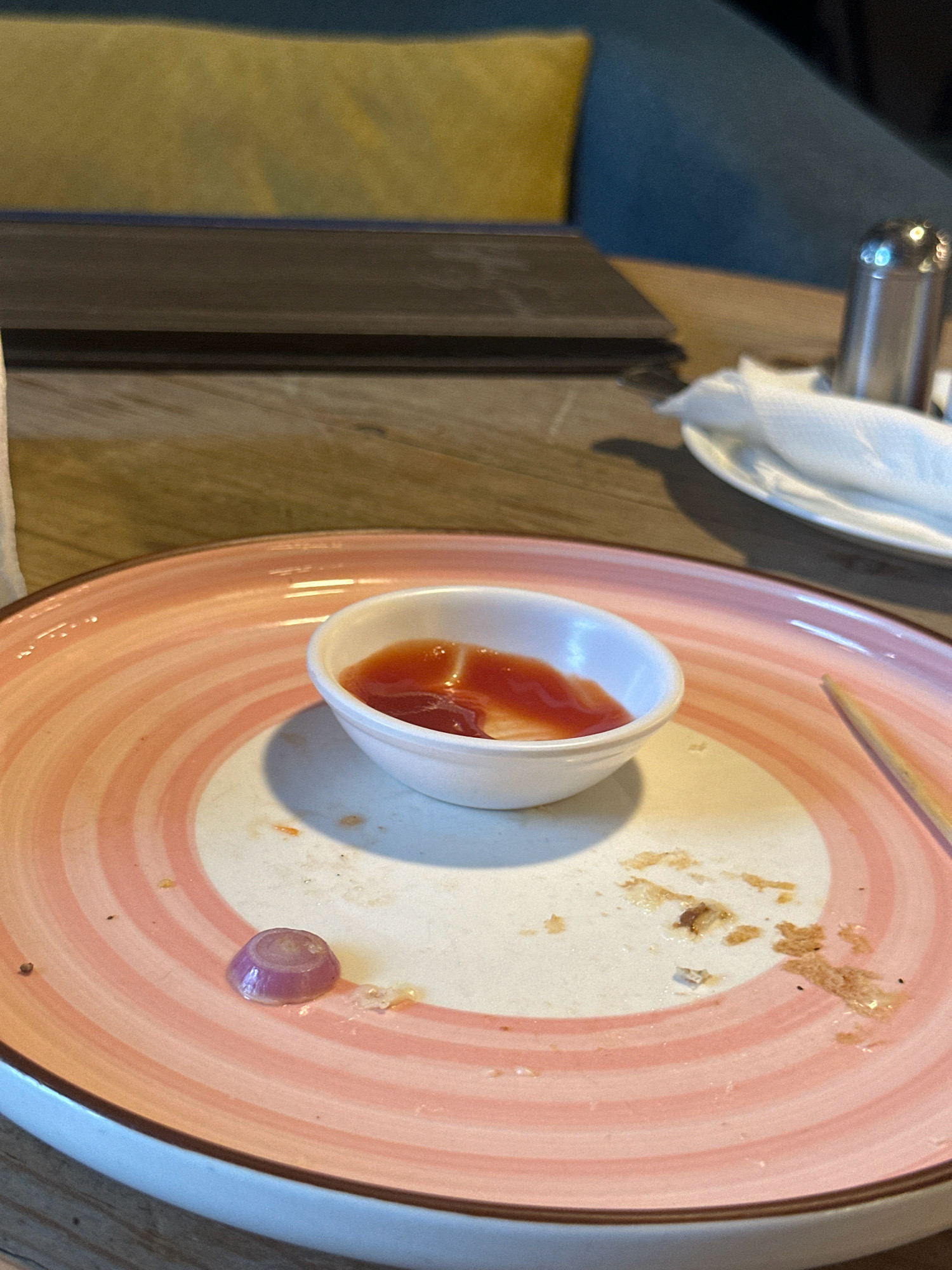 The image depicts a close-up of a pink and white swirled plate on a wooden table. On the plate, there is a small white ramekin containing ketchup. Scattered around the plate are remnants of food, including crumbs and a single slice of red onion. A napkin and a salt shaker are visible in the background. The lighting suggests an indoor setting, possibly a restaurant. The scene appears to be in a restaurant located in Kathmandu Metropolitan City, Nepal.