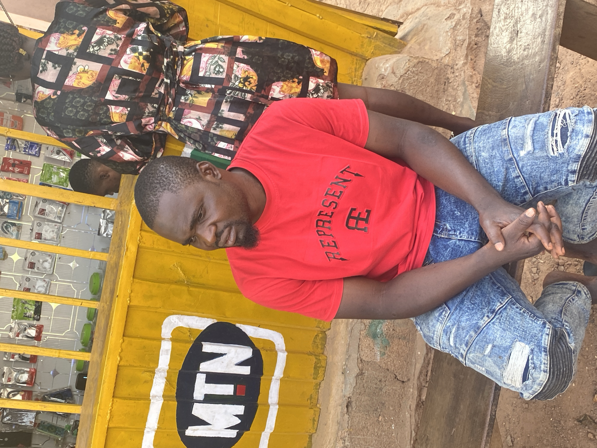 The image depicts an outdoor scene in Rido, Nigeria, centered around a yellow corrugated metal kiosk or stall. In the foreground, a dark-skinned adult male with short dark hair and a short beard is seated on a plain wooden bench, facing right. He wears a red short-sleeved t-shirt with "REPRESENT FE" printed in black, and distressed blue denim jeans with black ribbed knee panels. His hands are clasped together in his lap. Behind him, to the right, a person is partially visible from the back, wearing a long-sleeved patterned dress with a mix of black, red, yellow, and green geometric and floral designs. A second individual's head, with short dark hair, is visible further back and to the left of the patterned dress, obscured by the yellow structure. The yellow structure features a large "MTN" logo (white on a black oval with a white outline). Above the yellow structure, a display area shows various small packaged electronic accessories, such as cables and earphones, hanging from hooks against a light-colored background with a grid pattern. The ground appears to be packed reddish-brown dirt or earth. The scene is illuminated by natural daylight.