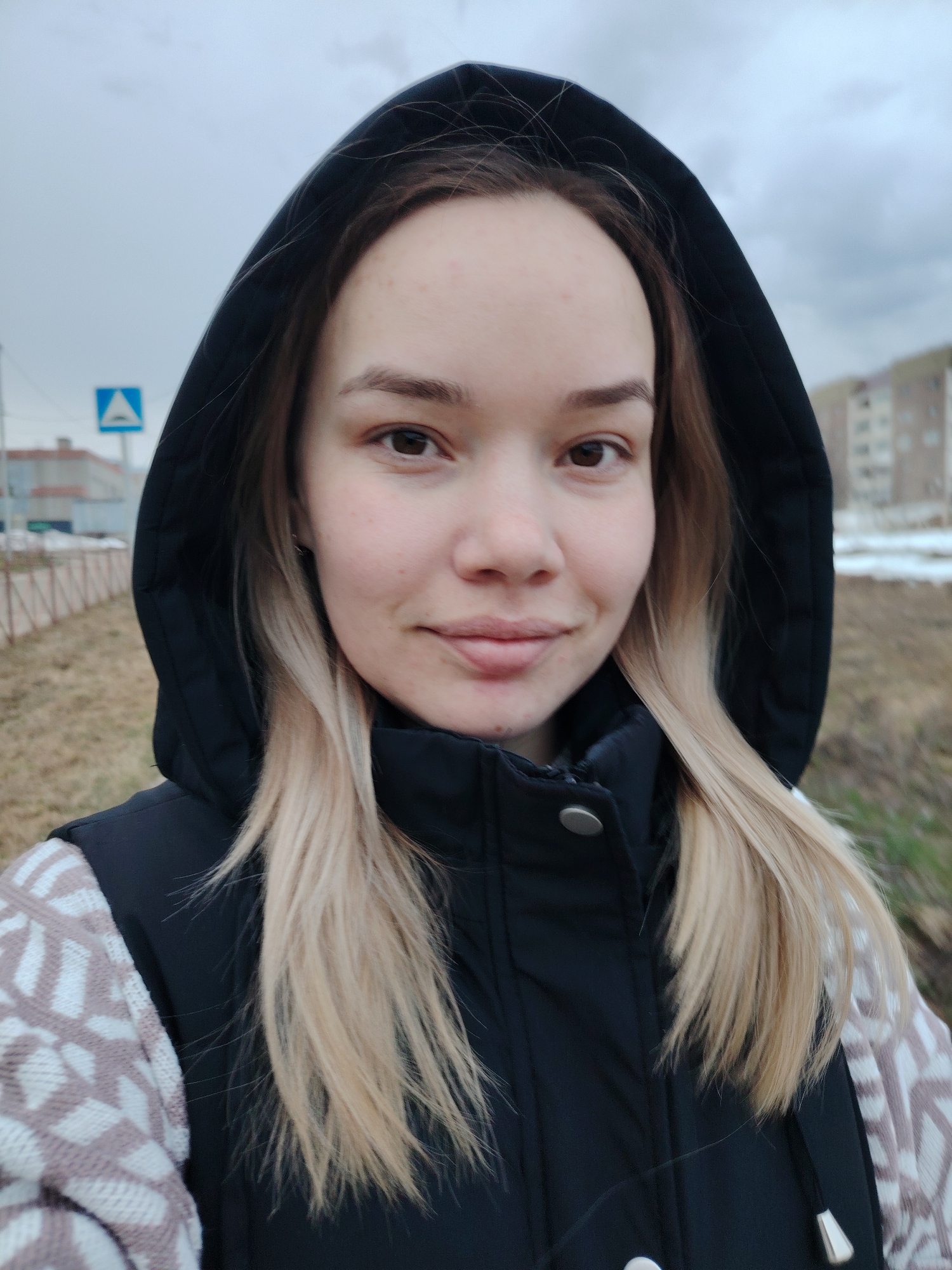 A young woman is standing outdoors, looking directly at the camera with a slight smile. She is wearing a dark-colored hooded jacket over a patterned sweater. The hood is up, framing her face. Her blonde hair falls on either side of her face.
Behind her, there's a street sign on a pole, a low fence, and a building in the background. Some patches of ground show sparse vegetation, with hints of snow or dead foliage. The sky is overcast.
The location is Markovskii, Russia.