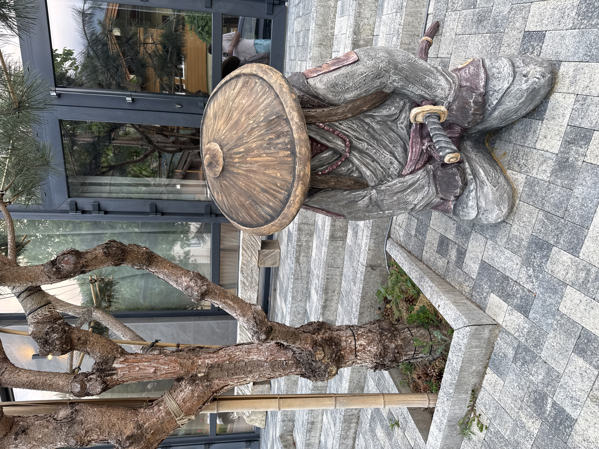 An outdoor scene in Krasnaya Polyana, Russia, features a stone statue of a samurai kneeling on a paved patio. The statue, wearing traditional samurai attire and a conical hat, holds a sword. To the left of the statue, a gnarled tree grows from a raised planter box filled with pinecones and greenery. Behind the statue and tree, concrete steps lead up to a building with large glass doors and windows. The paving stones are a mosaic of gray and white squares.