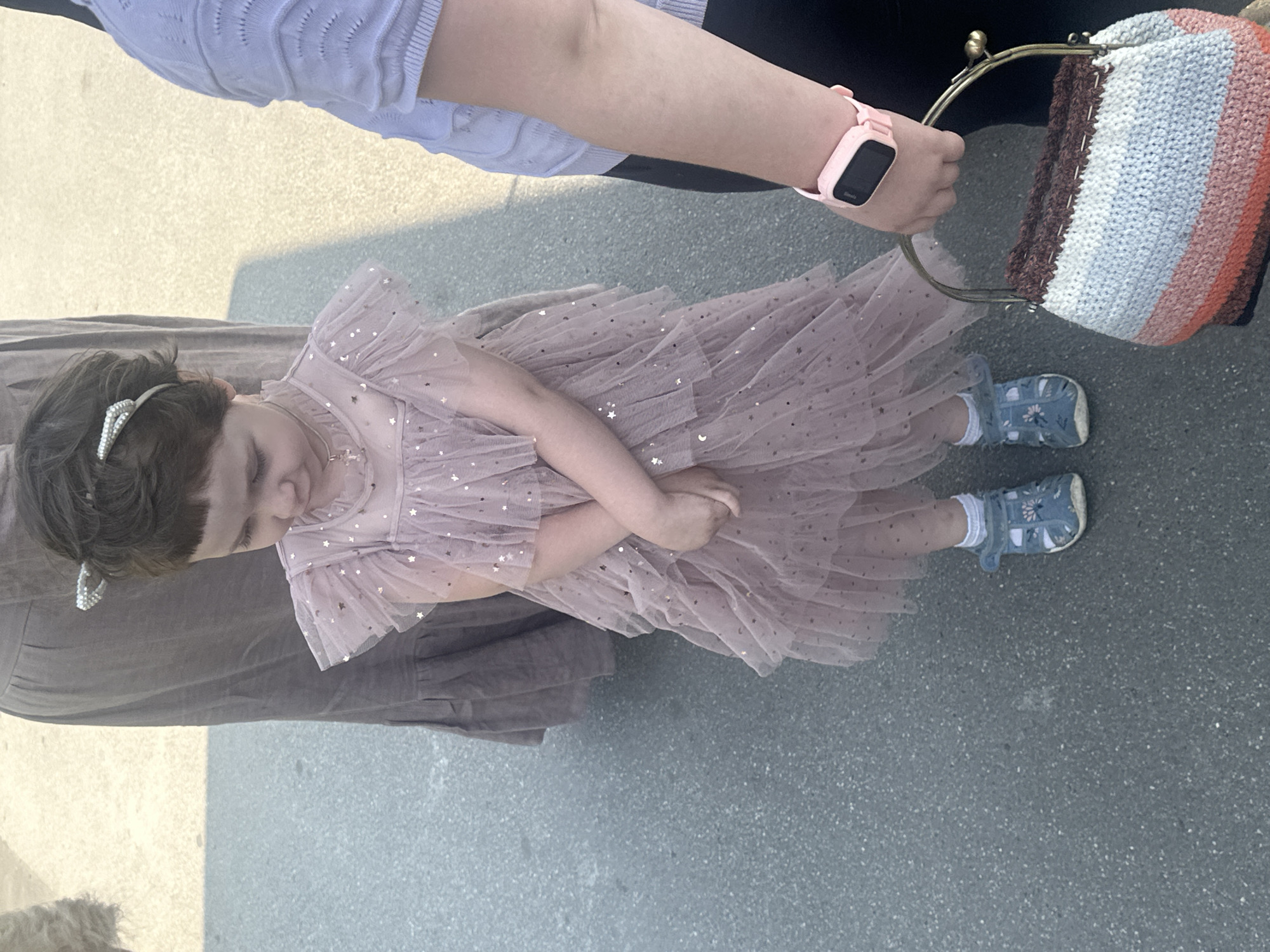 A young girl wearing a pink, sparkly dress and blue sandals stands on a paved surface. She has dark hair and is looking down. A woman's arm, wearing a pink smartwatch, is visible to the right of the girl, holding a crocheted bag with striped patterns in pink, white, and brown. Another person in a long, brown garment is partially visible to the left. The scene appears to be outdoors, possibly in a city, with a shadow cast on the ground from the right. The lighting suggests it is daytime.