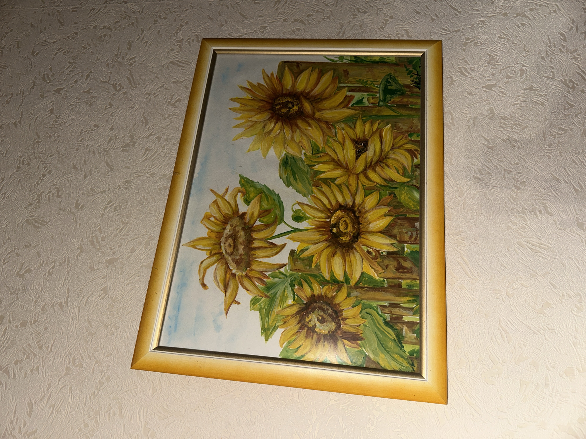 The image shows a framed painting of sunflowers hanging on a textured, light-colored wall. The painting depicts several sunflowers with yellow petals and brown centers, along with green leaves, against a background of a wooden fence and a pale blue sky. The frame is a golden color and appears to be slightly tilted. The lighting suggests the image was taken indoors, possibly in a room. There are no people visible in the image. The location is not specified beyond "Unknown, Unknown."