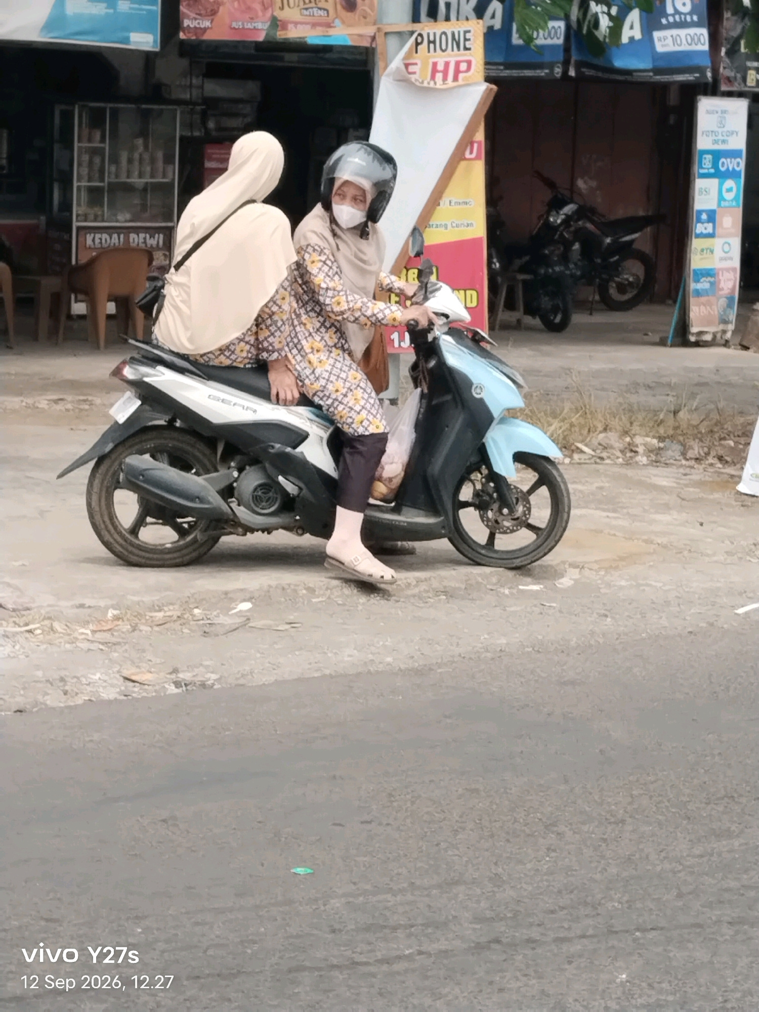 Two individuals are seated on a light blue and silver motorcycle on a paved road. The person in the rear, wearing a light beige headscarf and patterned shirt, is operating the vehicle. The passenger, also wearing a beige headscarf, is positioned behind the driver. Both appear to be wearing masks. The motorcycle has a license plate that is partially obscured. In the background, there are several shops and businesses. One shop displays various signs, including one with "PHONE" and "HP" on it, and another with "KEDAI DEWI" written on it. A black motorcycle is parked in front of one of the buildings. The overall environment suggests a commercial area within Trimulyo, Indonesia, likely a street scene with shops lining the road. The image is dated September 12, 2026, at 12:27.
