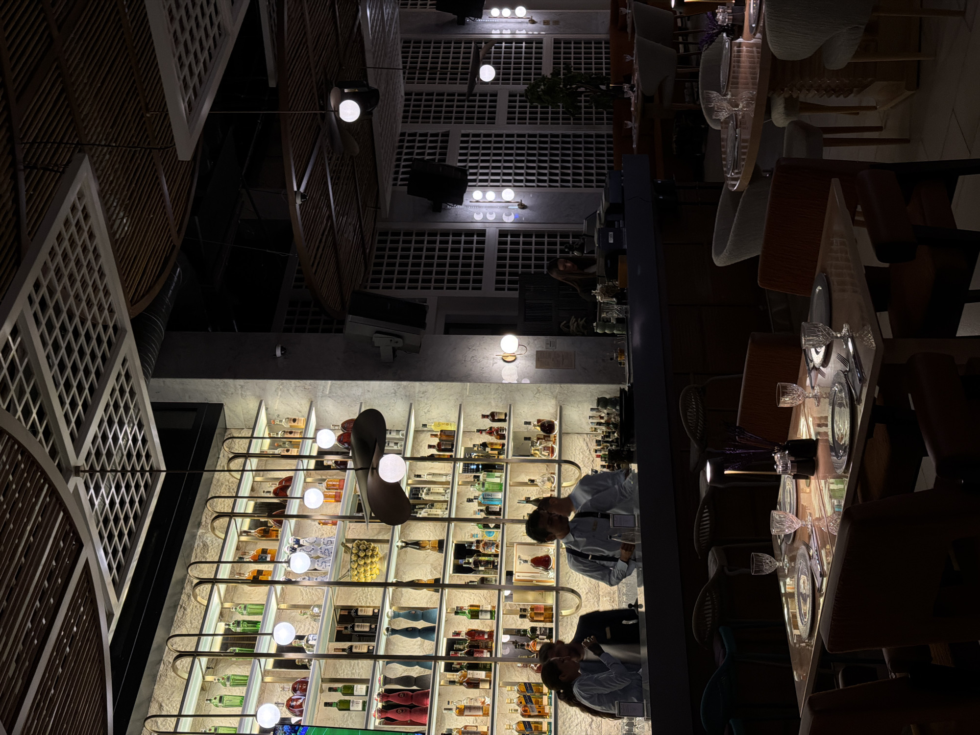 The image depicts the interior of a dimly lit, upscale bar and restaurant. The focal point is a well-stocked bar with shelves displaying numerous bottles of liquor and other beverages. Three individuals, likely staff members, are visible behind the bar. Two of them appear to be engaged in conversation or work, while a third is partially obscured. In the foreground, several tables are set for dining, complete with plates, cutlery, and glassware. The restaurant features modern decor, with patterned ceilings and ambient lighting. The overall atmosphere suggests a sophisticated dining and drinking establishment. The location is identified as Budva, Montenegro.