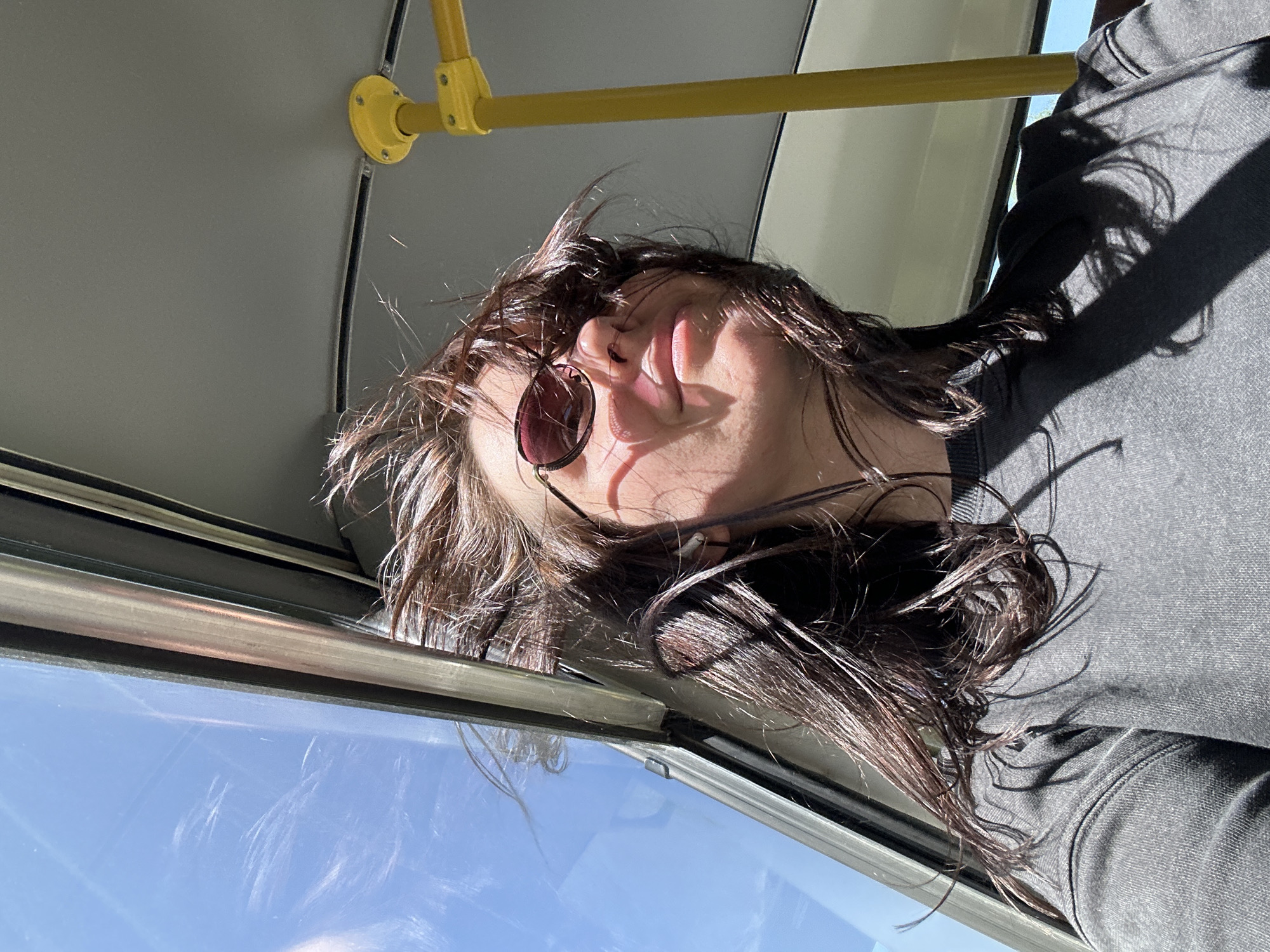 The image depicts a person wearing sunglasses with dark hair inside what appears to be a bus or public transportation vehicle. The person is looking up, possibly out of the window. The window shows a clear blue sky. A yellow handrail is visible on the ceiling. Based on the location provided, Ol'khovochka, Russia, the environment suggests an urban or suburban setting in that area.