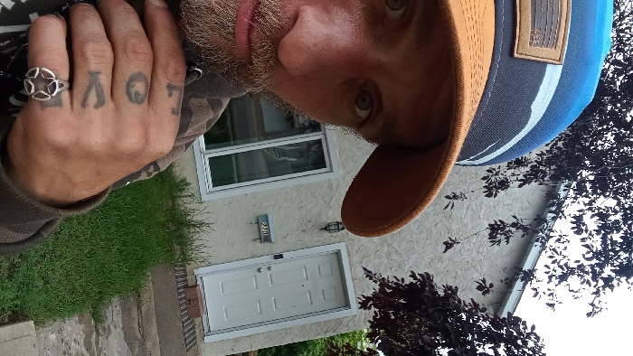 The image displays a male subject in a close-up, upward-angle selfie, positioned outdoors in front of a residential building identified by the municipal address "270" in Medicine Hat, Canada.
The subject's face, visible from the right eye to the nose and mouth, shows light-colored eyes, a light brown beard, and stubble. He is wearing a two-toned baseball cap with a blue crown, a brown brim, and a rectangular leather-like patch on the front. His attire includes a dark, possibly camouflage-patterned long-sleeved garment. The subject's right hand is prominent in the foreground, displaying knuckle tattoos that spell "LOVE" and a silver-colored ring with a five-point star design on the ring finger.
The background consists of a single-story or lower unit of a light beige, stucco-textured building with white trim. A white six-panel entry door is visible on the right, with a dark rectangular doormat and a small brown cardboard box resting on the concrete stoop. To the left of the door, a black, wall-mounted lantern-style light fixture is installed above a light blue rectangular plaque displaying the numerals "270." A large, white-framed window with reflective glass is situated to the left of the entrance. The ground immediately in front of the structure is covered in green grass of varying height, and a concrete pathway is visible in the foreground. Dark-leaved deciduous trees or shrubs partially obscure the building's upper and side portions. The sky above is uniformly overcast, indicating diffused natural light conditions.