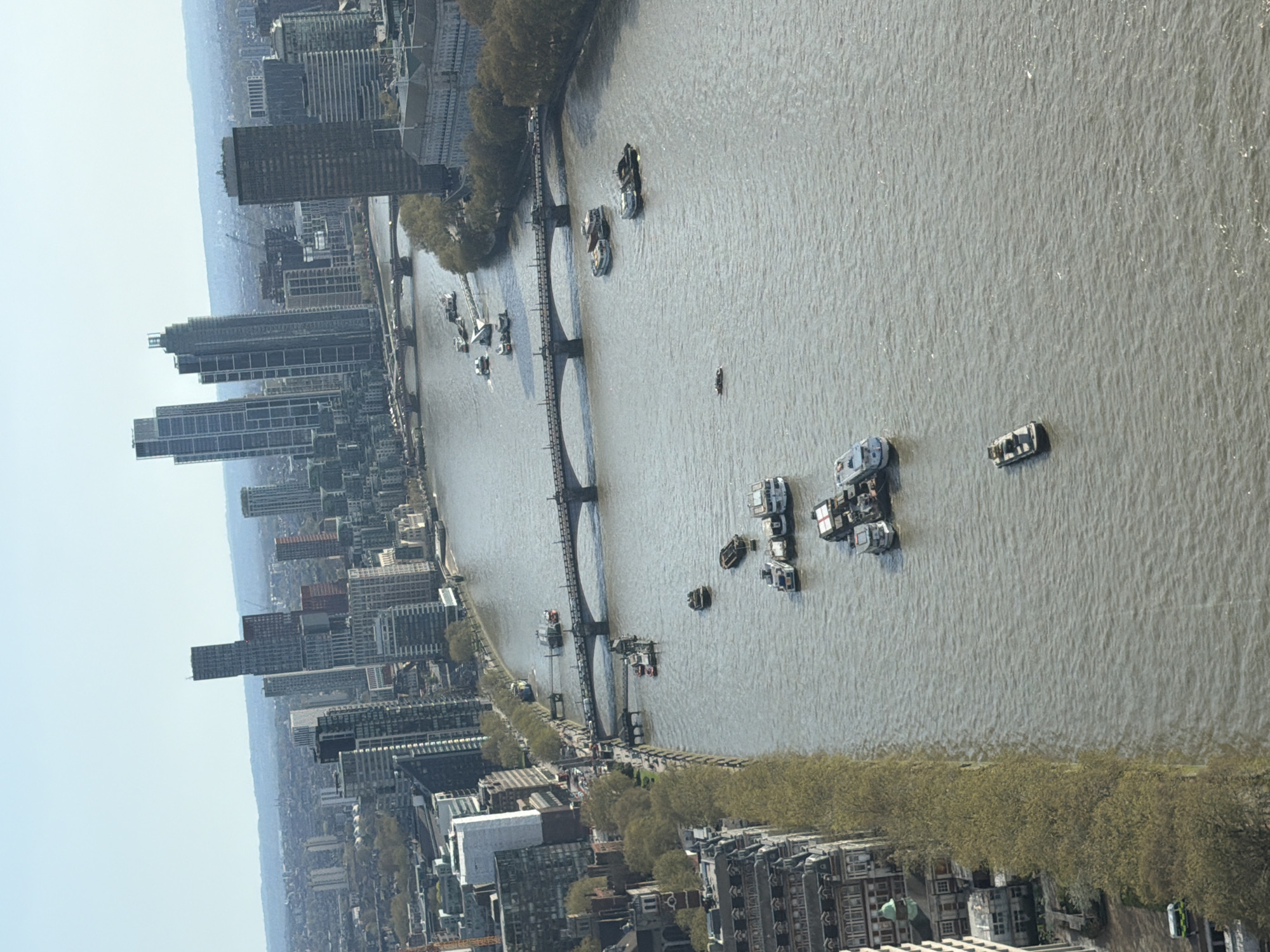 The image shows an elevated view of the River Thames in London, United Kingdom. A bridge with multiple arches spans the river, connecting the cityscape on both sides. Numerous boats and other watercraft are seen docked or moving along the river. Buildings of varying heights and architectural styles, including modern high-rise apartments and older structures, line the riverbanks. Trees border the river's edge. Pedestrians appear to be walking along the embankment. The sky is bright with few clouds. The river's surface is textured, indicating possible light wind or current.