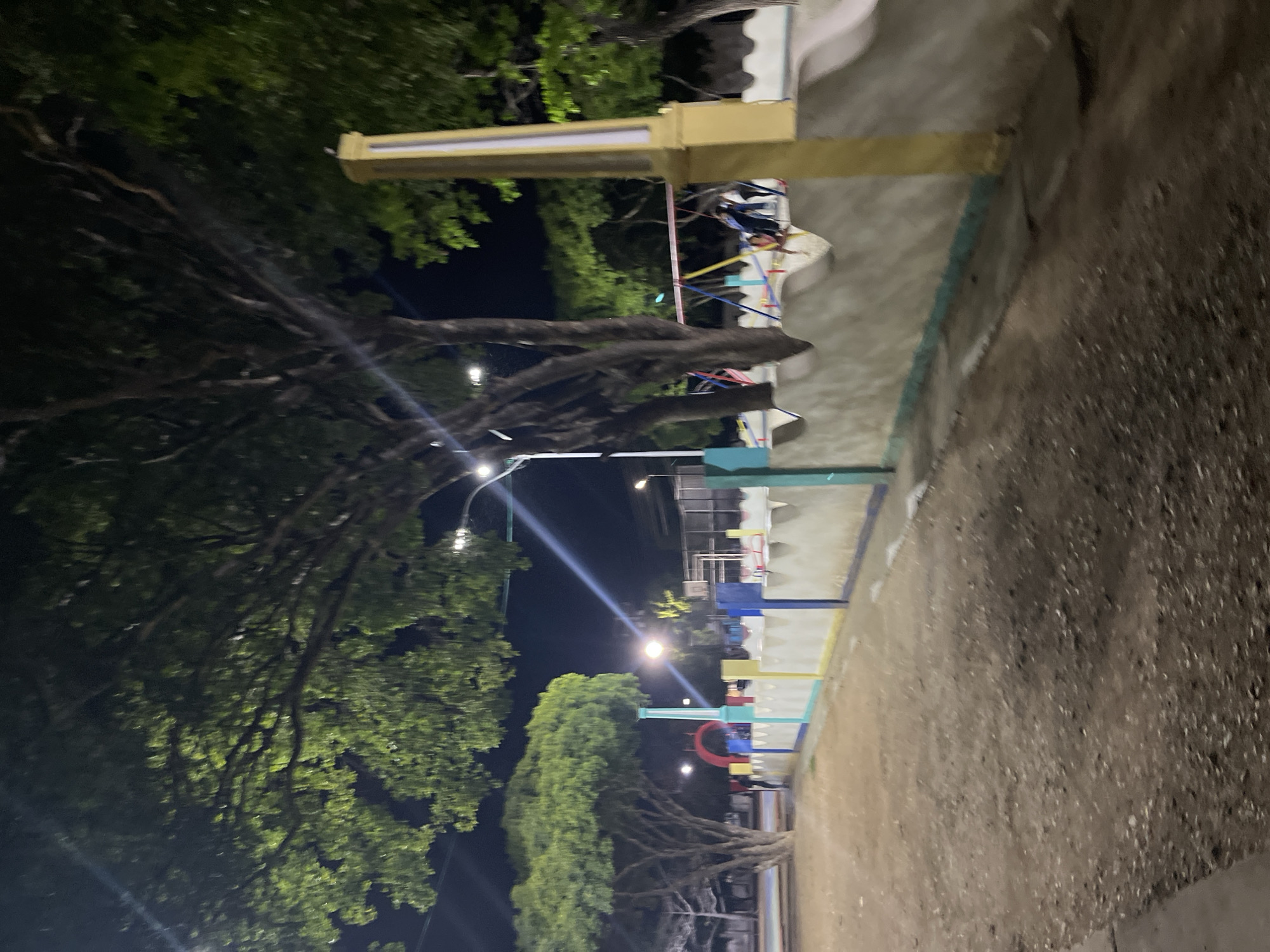 The image captures an outdoor scene at night, likely a public park or recreational area, in Santa Ana, Venezuela. A low, decorative wall, painted with colorful bands of yellow, blue, and green, runs along the foreground. Beyond the wall, a playground area is visible, with swings and some structures that suggest playground equipment. Several trees with dense foliage are prominent throughout the scene, some illuminated by artificial lights, creating strong shadows. The ground appears to be dirt or gravel. In the background, there are hints of buildings and sports courts, possibly a basketball court. A few individuals are present in the playground area, though their actions are not clearly discernible. The overall impression is a dimly lit public space, potentially a community gathering spot or park, in Santa Ana.