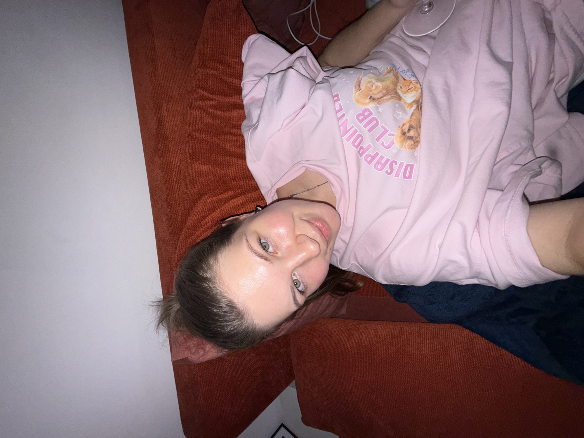 A female subject is depicted in a high-angle selfie photograph, lying on an orange, textured fabric sofa with cushions in an indoor setting within the city of Vozdovac Urban Municipality, Serbia. Her head is positioned on a cushion, with her brown hair gathered into a high bun/ponytail. She has fair skin, green eyes, and displays a slight smile while looking directly into the camera. She is wearing a light pink, short-sleeved t-shirt featuring the text "DISAPPOINTED CLUB" in pink lettering, accompanied by an image of three light brown rabbits/bunnies. A silver-colored chain necklace is visible around her neck. A dark blue blanket or throw is partially visible beneath her left side. The background consists of a white wall, with a portion of a framed object visible on the upper left. A small, circular, clear object with a white rim is situated on her t-shirt near her right arm. The scene is illuminated by an interior light source.