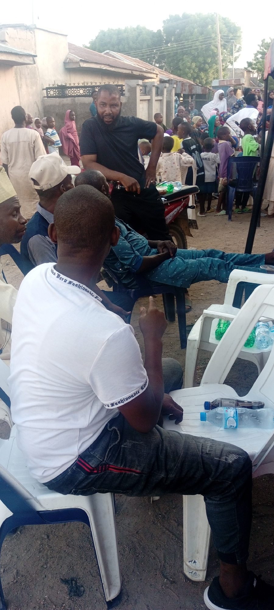 This image, captured in Gwagwarwa, Nigeria, depicts an outdoor gathering featuring several adults in the foreground and a crowd of individuals, predominantly children, in the background. In the immediate foreground, from left to right, an adult male is partially visible, wearing a light-colored cap. Adjacent to him, another adult male is seated with his back to the camera, wearing a white short-sleeved t-shirt with "Very Fashionable" text along the sleeves, dark jeans with red side stripes, and black sneakers; his right index finger is raised. To his right, a third adult male, also seated and viewed from behind, wears a dark blue polo shirt and a white baseball cap. A fourth adult male, with a dark beard and wearing a black short-sleeved t-shirt and dark pants, leans forward, resting his arm on a surface, and looks directly at the camera. Behind him, a fifth adult male, dressed in a light blue patterned shirt and matching pants, sits with his legs extended and feet resting on an unseen object.
On a white plastic chair to the right of the man in the white t-shirt, two plastic bottles (one with a dark liquid, one with clear water) and a clear plastic bag containing green items are present. A red motorcycle with a black seat and a white plastic bag on it is partially visible behind the man in the black shirt.
In the midground and background, a diverse group of people, mostly children dressed in various casual attire, are gathered. Visible among them are a child in a pink garment, an adult woman in a white hijab, and an adult carrying a baby. Behind this crowd, structures with light-colored walls and corrugated metal roofs are present, along with green trees. The ground is composed of dirt, and the lighting indicates a late afternoon or early evening setting.
