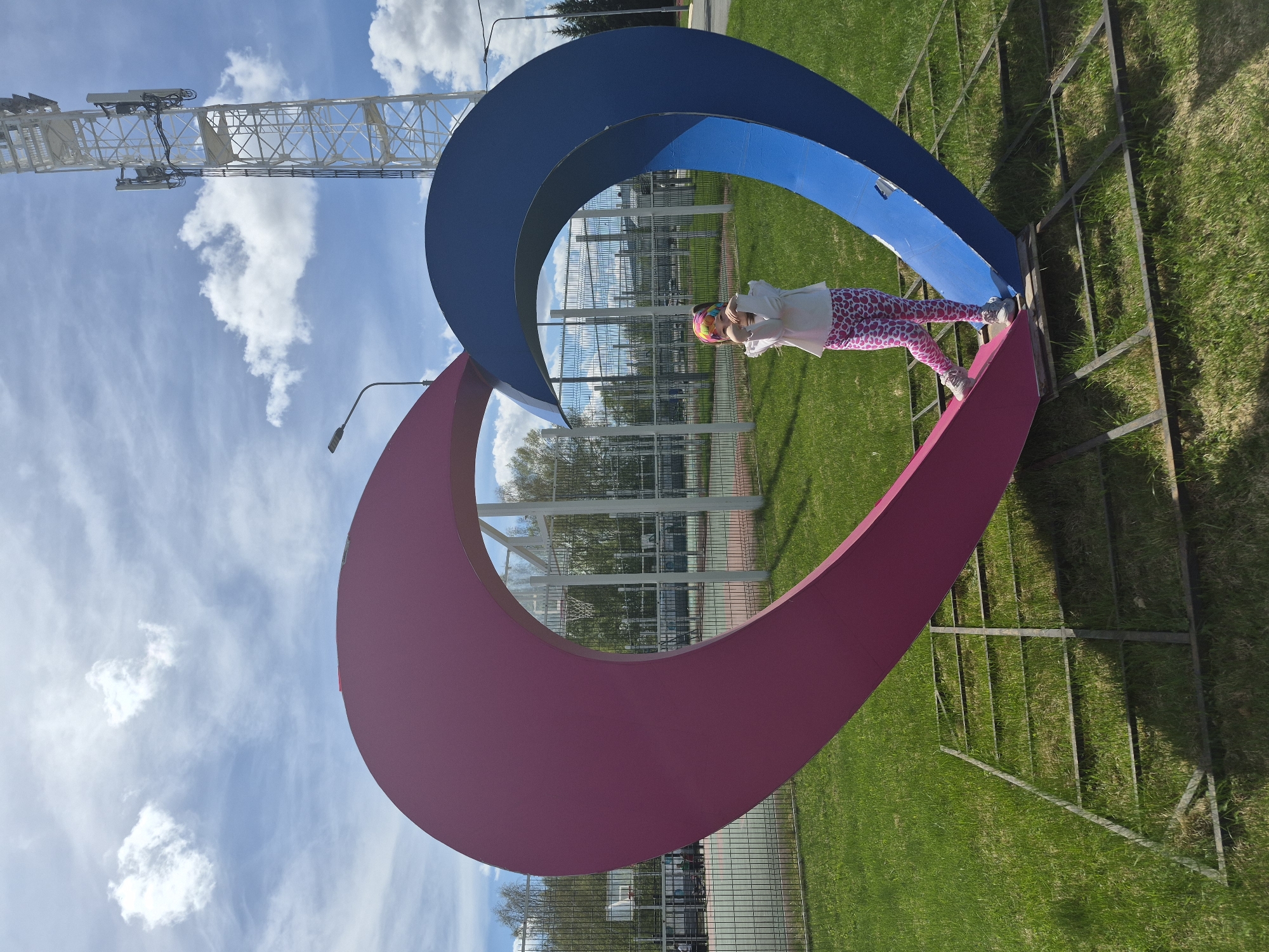 The photograph depicts a young child positioned on a large, abstract heart-shaped sculpture in what appears to be an outdoor public space. The sculpture is constructed from broad, curved metal pieces painted in blue and pink. A light tower is behind the sculpture and in the background of the frame. The child is wearing a light-colored long-sleeve shirt, spotted pink leggings, and a multicolored headband. They are resting their hands on the blue portion of the sculpture. The background includes green grass, a metal fence, trees, and a partial view of buildings. The sky is blue with scattered clouds. Based on the style of the metal fence and the architecture of the visible buildings, the photograph was taken in the city of Tobolsk, Russia.