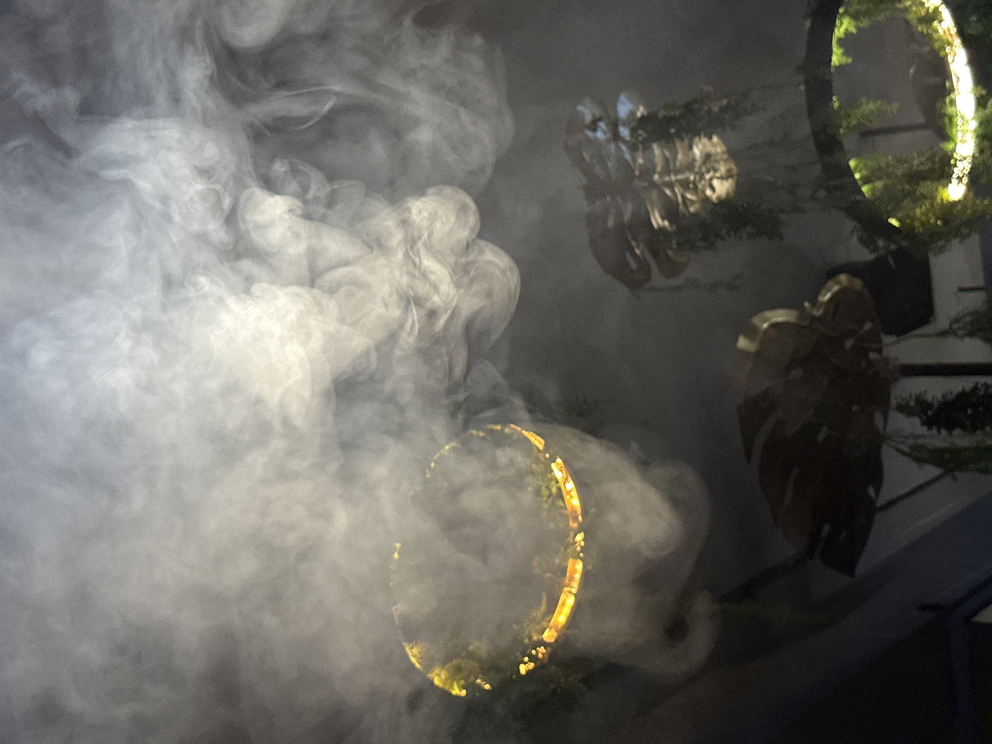In a dimly lit environment, possibly a bar or restaurant in North Kuta, Indonesia, a dramatic scene unfolds with abundant white smoke or fog creating an ethereal atmosphere. Two circular, illuminated hanging decorations are visible, each adorned with lush greenery, possibly hanging plants or moss. One decoration, closer to the foreground and on the left, is prominently engulfed in the dense smoke, its golden inner light shining through. Another, slightly further back and to the right, is partially obscured by the smoke, but its intricate structure and suspended greenery are discernible. The overall impression is one of mystery and allure, with the smoke acting as a veil, obscuring and highlighting the decorative elements and creating a theatrical ambiance.