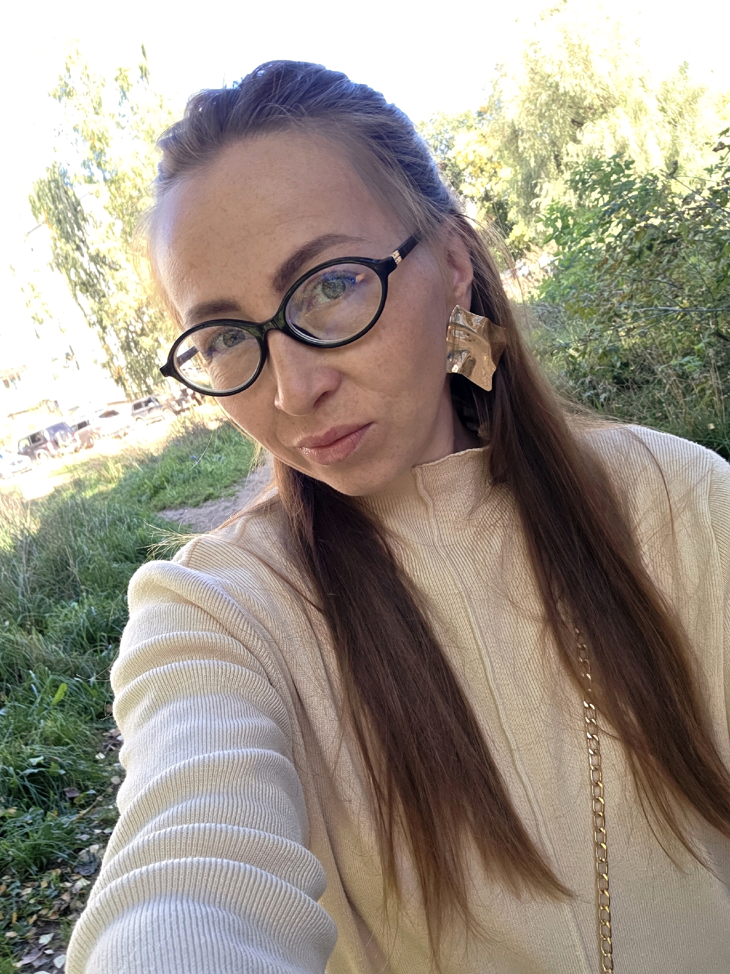 A medium shot is presented of a woman wearing black-rimmed glasses and a cream-colored sweater. She has long, brown hair that falls over her shoulders and is wearing large, gold, abstract earrings. The woman is outdoors, with a background of green grass, fallen leaves, and trees. In the distance, several cars are parked on a paved surface. The lighting suggests it is daytime, possibly during autumn. The specific location within Chaikovsky, Russia is not discernible from the image.
