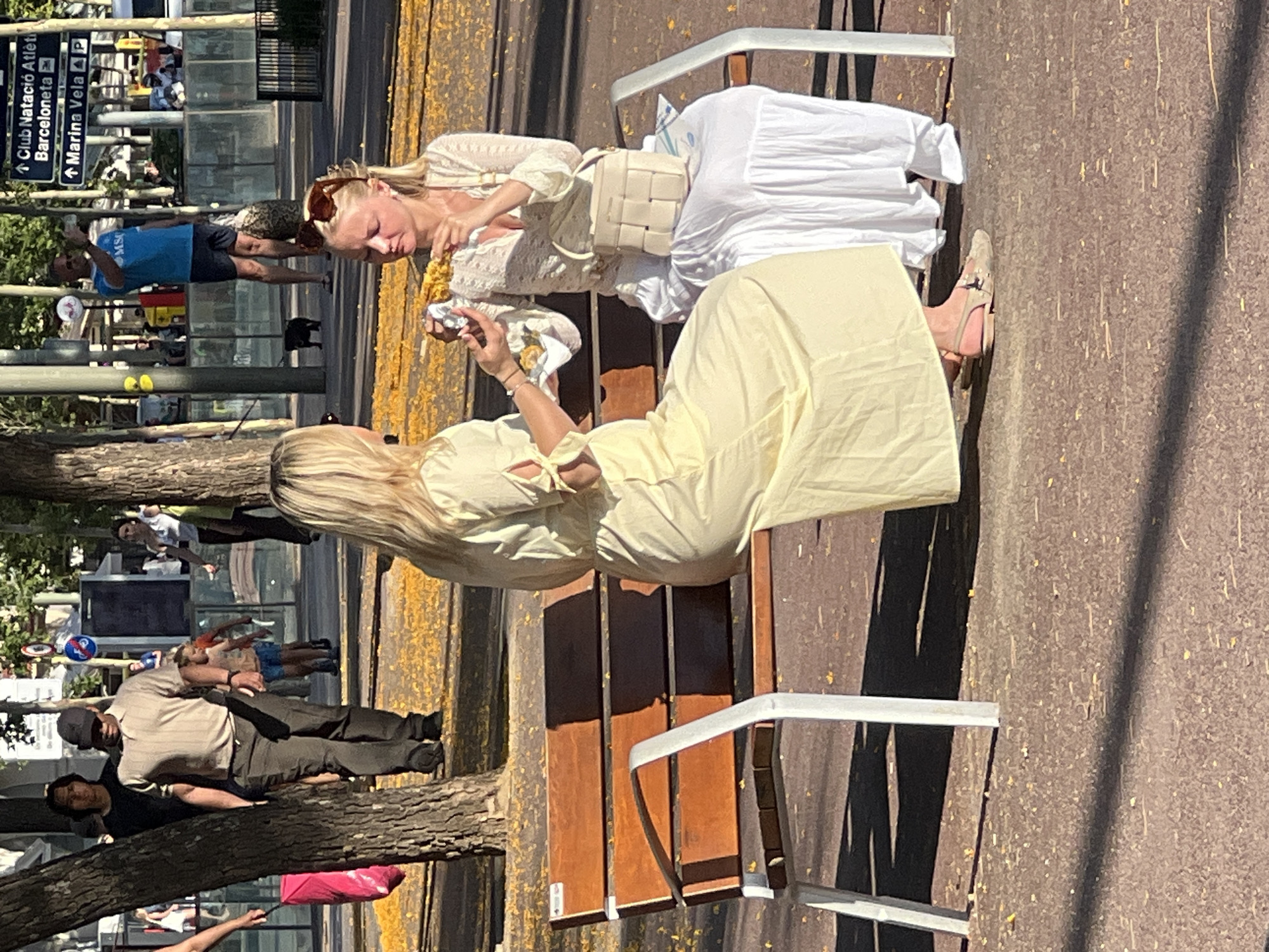 This image depicts a sunlit urban scene in Barcelona, Spain, identified by a street sign pointing to "Club Natació Atlètic Barcelona" and "Marina Vela P." In the foreground, two women are seated on a brown slatted public bench with light grey metal supports. The ground is paved and scattered with numerous small, yellow fallen leaves or blossoms.
The woman on the left, with blonde hair, wears a light yellow, long-sleeved maxi dress with tie details on the back of the sleeves. She is turned towards the woman on the right, holding a small silver foil-wrapped object in her right hand, appearing to offer or handle food. She wears light-colored flat shoes.
The woman on the right, also with blonde hair and sunglasses pushed onto her head, wears a light-colored, possibly cream, long-sleeved, open-knit top and a long, flowing white skirt. She holds and is focused on eating a yellow, possibly corn-on-the-cob-like food item. A small, light-colored, woven handbag rests on the bench beside her.
In the mid-ground, a large tree trunk is prominent on the left. Behind it, a man in a light brown polo shirt and dark pants, wearing a dark baseball cap, walks towards the right. Further back, a man in a blue t-shirt with "MSG" visible, wearing shorts, stands near a pole. He is holding a phone, possibly taking a picture, and has a patterned bag at his feet with a small black dog on a leash. Direct sunlight casts distinct shadows from the people, trees, and bench. Beyond the figures, a street and other urban structures are visible.